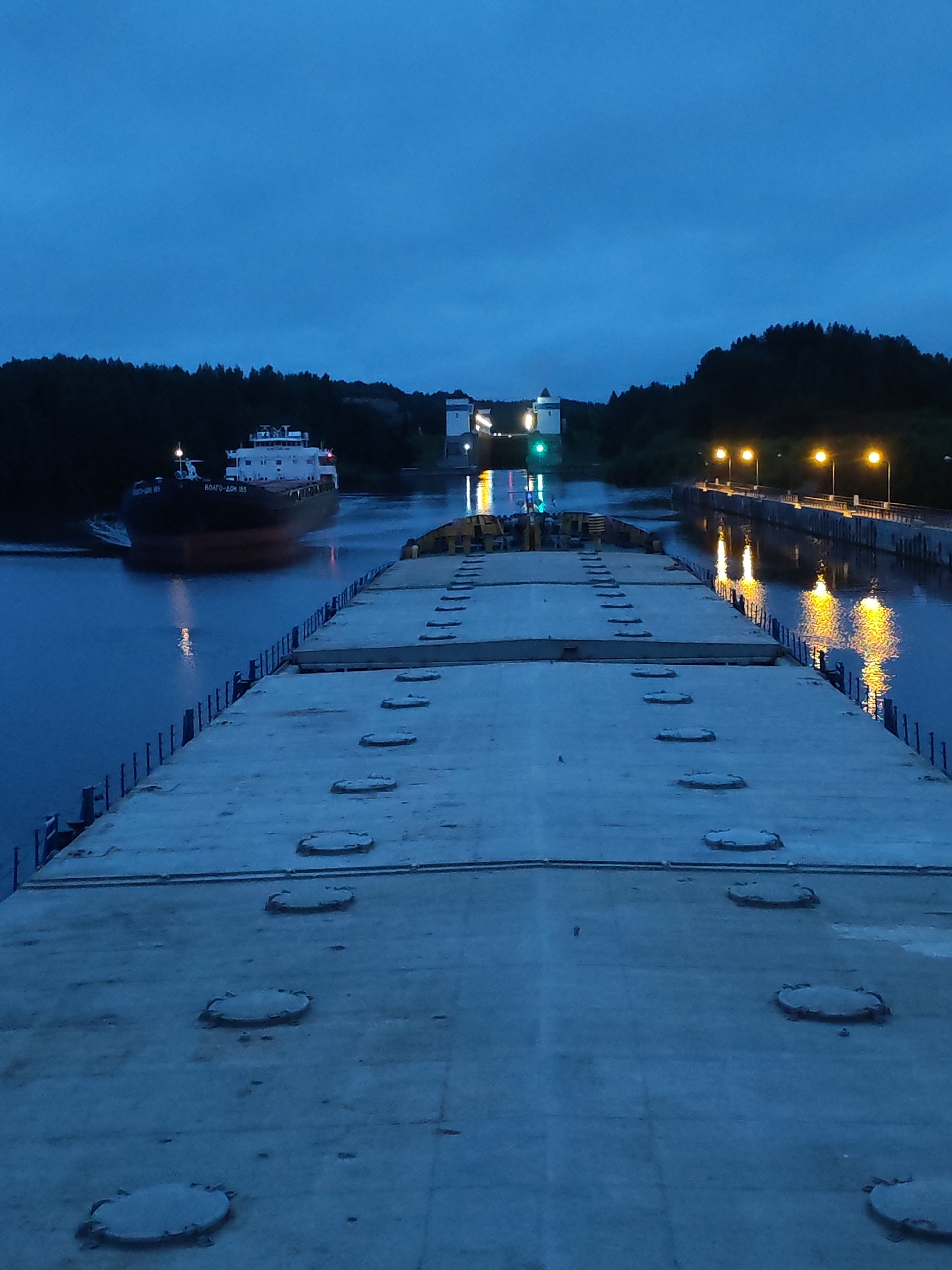 A cargo ship is visible in the water at dusk, approaching a lock system. The ship is dark with a white superstructure and appears to be a large cargo vessel. In the foreground, the perspective is from the deck of another, larger vessel or structure, which is made of concrete and has several circular hatches visible. The water reflects the lights from the lock system and the cargo ship. The background shows a treeline and the lock gates, with lights illuminating the area. The sky is a deep blue, indicating twilight. The location is identified as Markovo, Russia.