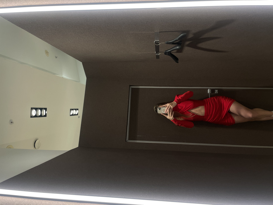 A woman in a short red dress is reflected in a mirror within a changing room. She is holding a phone, presumably taking a photo of herself in the mirror. The changing room is mostly brown with a white corner with lights. A clothes hanger is visible above the woman in the mirror. Fluorescent lighting is present at the top and bottom of the image frame.