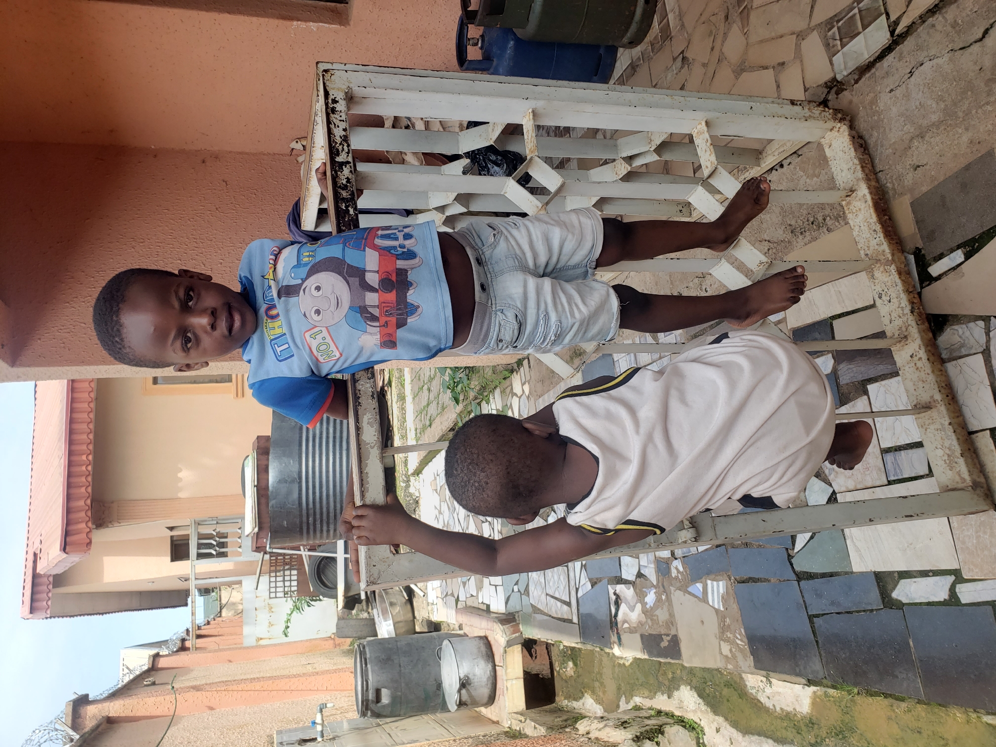 Two young boys are in an outdoor courtyard in Sabon Tasha, Nigeria. The boy on the right, facing forward, is standing and wearing a blue t-shirt with a Thomas the Tank Engine graphic and light denim shorts. He is leaning against a white metal gate. The boy on the left, with his back to the camera, is hanging from the same gate by his hands, with his feet dangling. He is wearing a light-colored tank top. The courtyard has a tiled floor and a drain on the left side. In the background, there are buildings, a large metal tank, and gas cylinders. The overall scene suggests a residential area.