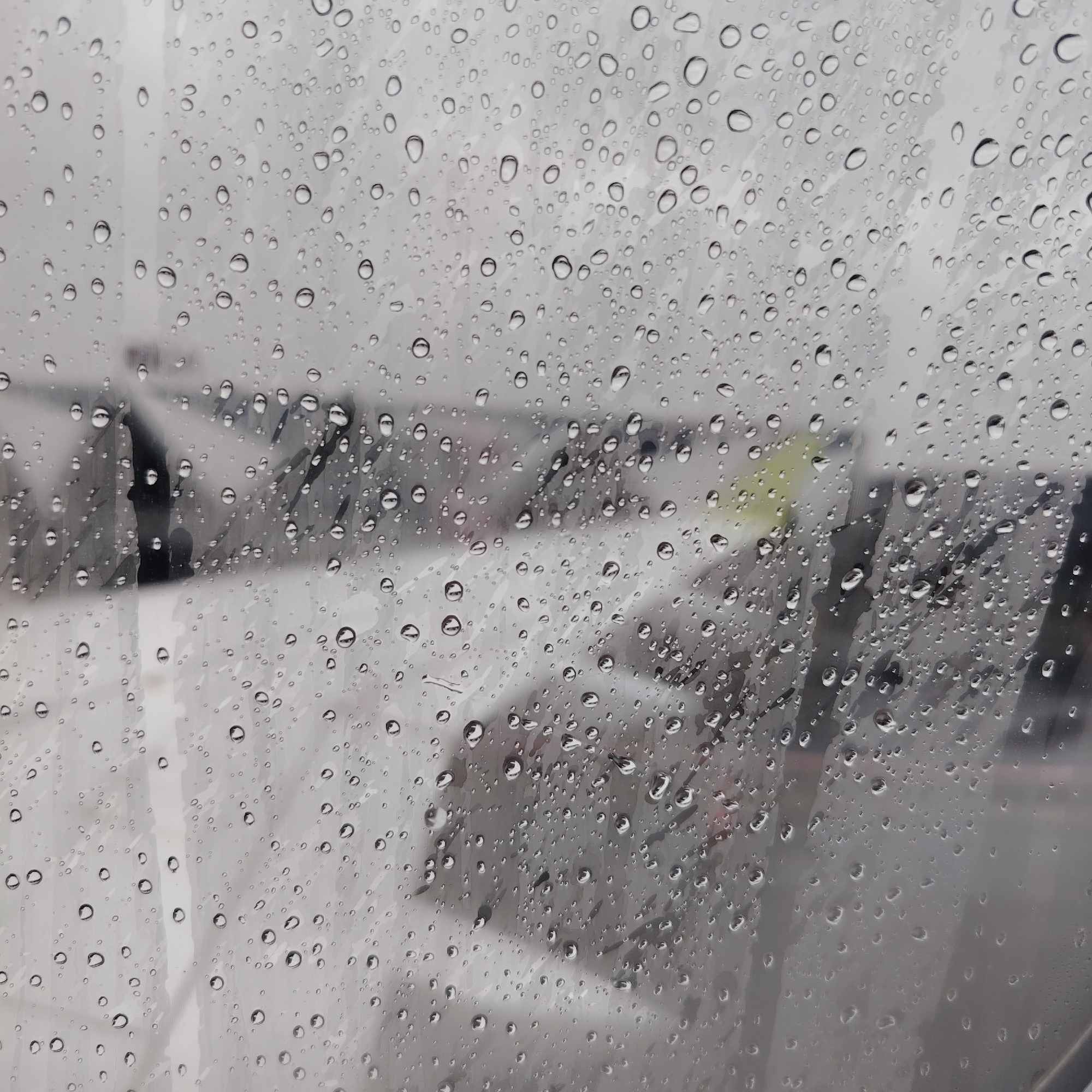 The image presents a close-up view through a transparent surface heavily coated with numerous water droplets of varying sizes. These clear raindrops refract and distort the out-of-focus background. The obscured background primarily consists of diffused white and light grey tones, indicative of a snow-covered or overcast outdoor environment. Indistinct, dark, vertical, and diagonal elements, consistent with bare tree branches or structural components, are vaguely discernible throughout the mid-ground. A subtle, diffused yellow-green tint is present in the upper-right quadrant of the background. No specific individuals, clearly defined objects, or discernible actions are visible beyond the environmental features. The scene is located in Skulte, Latvia, and suggests inclement, wet weather conditions.