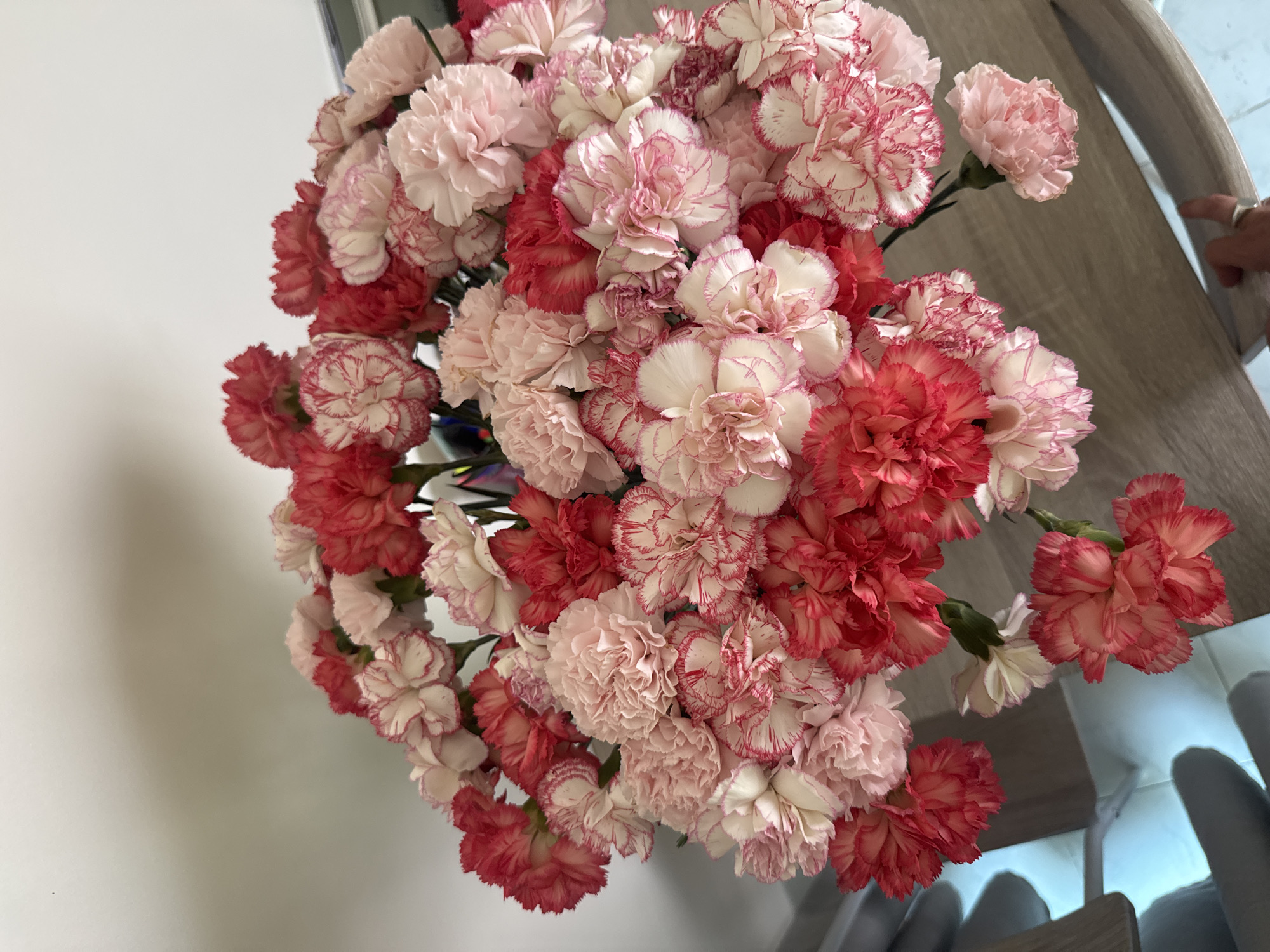 A dense floral arrangement, composed of numerous carnations in shades of solid pink, solid red, and bi-colored white with red margins, occupies the central and upper portions of the image. The flowers rest on a light-toned wooden surface, possibly a table or furniture top. A plain, light-colored wall is visible in the background. In the lower right corner, a human hand, wearing a silver-toned ring on the ring finger, is partially visible and in contact with the wooden surface. The scene is located in Choeng Thale, Thailand.
