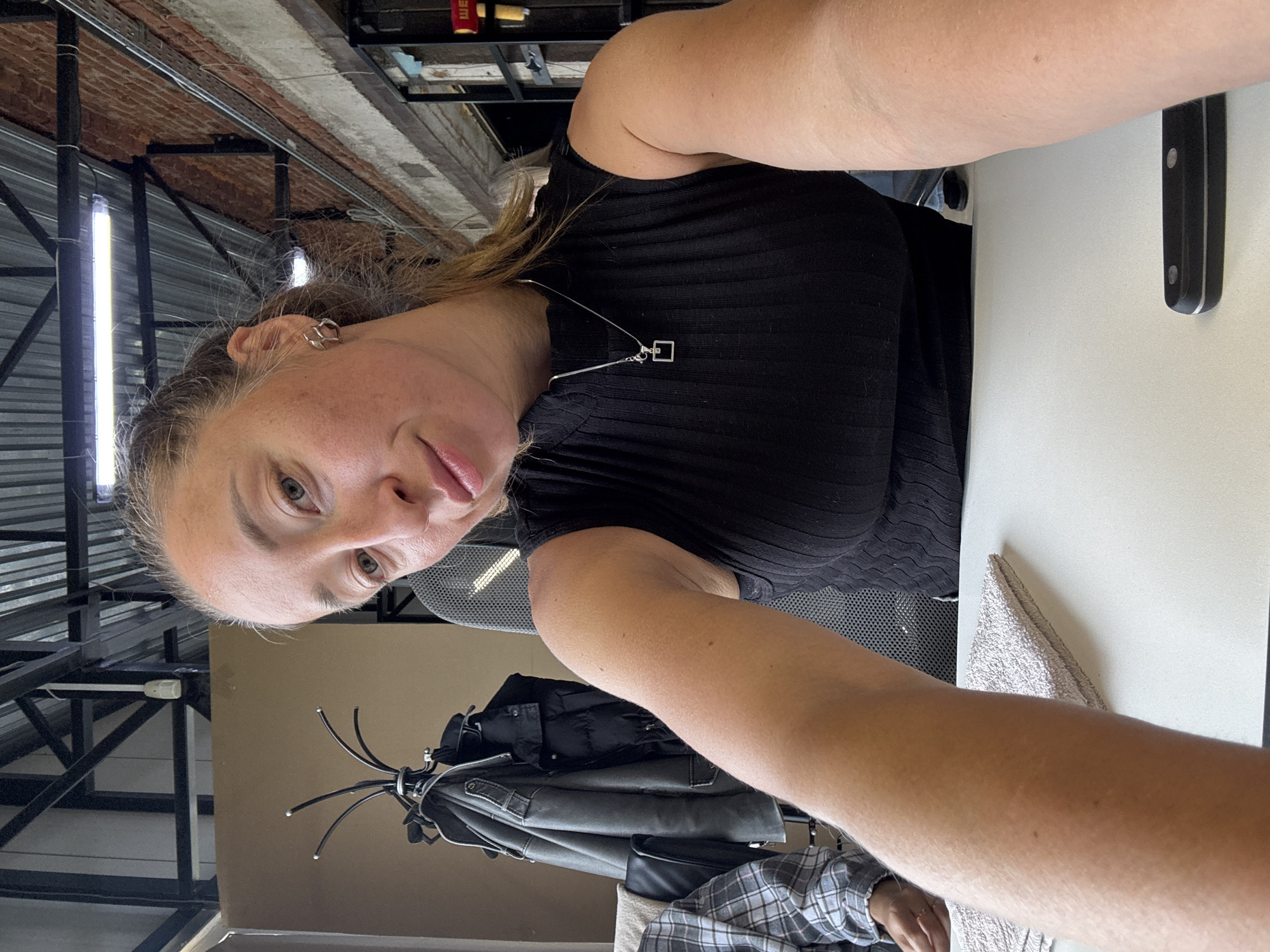 The image appears to be a selfie taken indoors. A woman with light skin, fair hair, and green eyes is the primary subject. She is wearing a black top and a silver necklace with a square pendant. An earring is visible in her left ear.
In the background, there is a clothes rack with several jackets hanging on it. There's also what appears to be a gray and white plaid shirt. To the right, there is a white table with a knife on it, and a light colored towel. The architectural features suggest an industrial or renovated space, with exposed metal beams and brickwork on the ceiling. Long, vertical lights are also visible.
While the image offers details about the interior, there is no specific information to definitively determine its location as Chaikovsky, Russia. Therefore, confirming the city of Chaikovsky, Russia as the location is not possible based solely on the image.