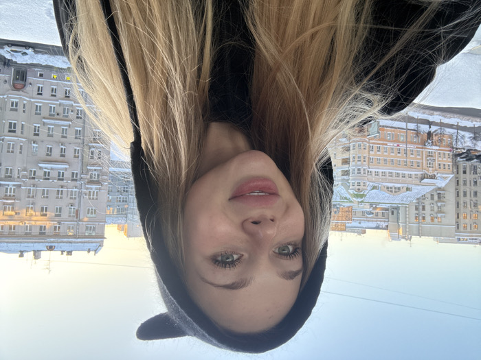 A front-facing close-up photograph depicts a young female subject indoors, capturing a selfie. The female has long blonde hair, blue-green eyes, and a light complexion, with her lips slightly parted in a faint smile. She wears a black hooded garment with two prominent ear-like protrusions on the hood. The background reveals a dense cityscape from an elevated perspective, featuring multiple large, multi-story buildings, some with ornate architectural details and snow-covered roofs. Notably, a light orange/beige building with a distinctive green/grey patterned roof is visible in the mid-ground left. A street below is partially visible, also covered in snow, with some vehicles present. The sky is clear, transitioning from light blue to pale yellow near the horizon, indicative of either sunrise or sunset. Power lines are faintly visible against the sky. No other individuals or specific interactions are discernible. The environment suggests a winter season in an unidentified urban area, referred to as Unknown, Unknown.