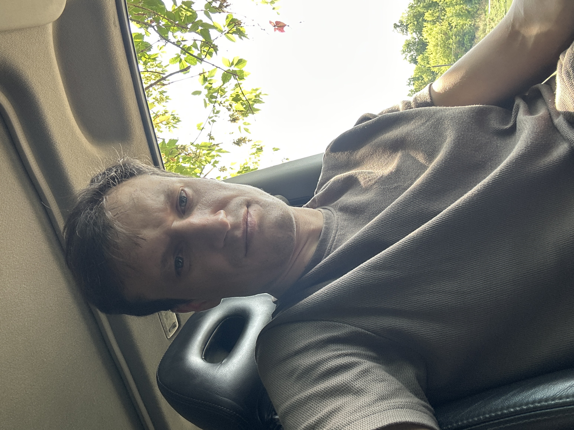 A close-up selfie of a man sitting in the passenger seat of a car is depicted. The man has light skin, short brown hair, and is looking directly at the camera with a neutral expression. He is wearing a brown t-shirt. The interior of the car is visible, including a black leather headrest and the car's ceiling. Through the car window, lush green trees and a bright sky are visible, suggesting the car is outdoors in a natural environment. The image is well-lit, with sunlight filtering through the leaves of the trees. The provided location of Makiv, Ukraine, places this scene in a rural or suburban setting within that region.