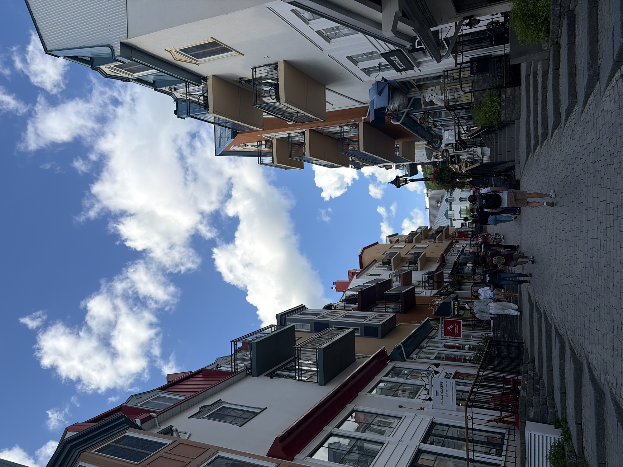 The image depicts a view of a street in Mont-Tremblant, Canada, with multi-story buildings lining both sides and a cobblestone street leading uphill. The buildings feature a mix of architectural styles, with prominent balconies, red roofs, and colorful facades. Several people are walking up the street, some carrying backpacks. Shops with signs such as "RUDSAK" and "ENGEL & VÖLKERS" are visible at street level. The sky is blue with scattered white clouds.