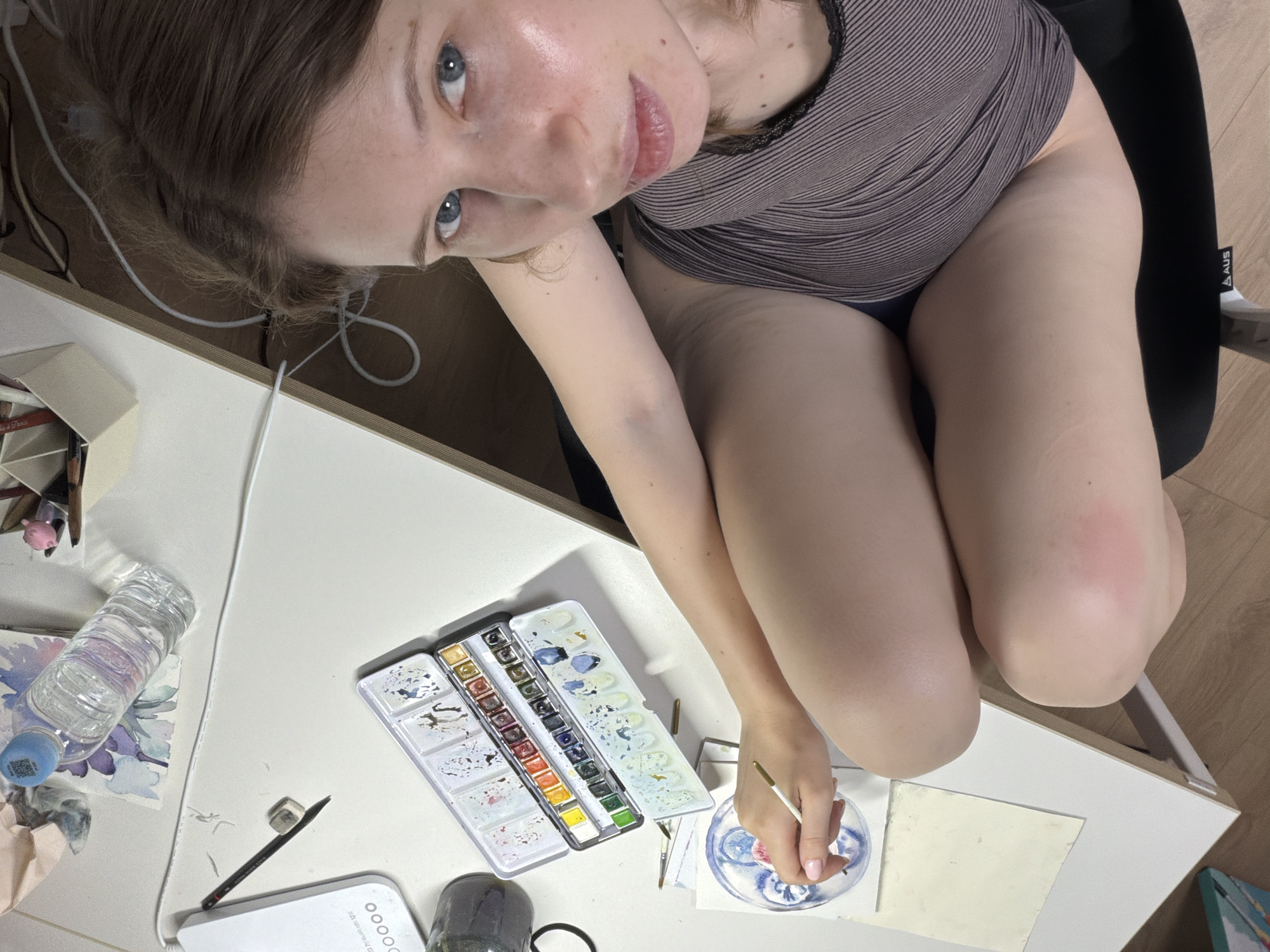 The image shows a woman with light skin and blue eyes, her hair is light brown. She is wearing a striped tank top and appears to be sitting at a desk. She is holding a paintbrush in her right hand and appears to be painting on a small piece of paper. On the desk, there is a watercolor paint set, a bottle of water, and other art supplies. The desk is white and appears to be in a room with wooden flooring. The overall lighting suggests it is daytime. The context of Dongducheon-si, South Korea is not visually ascertainable from the image itself.