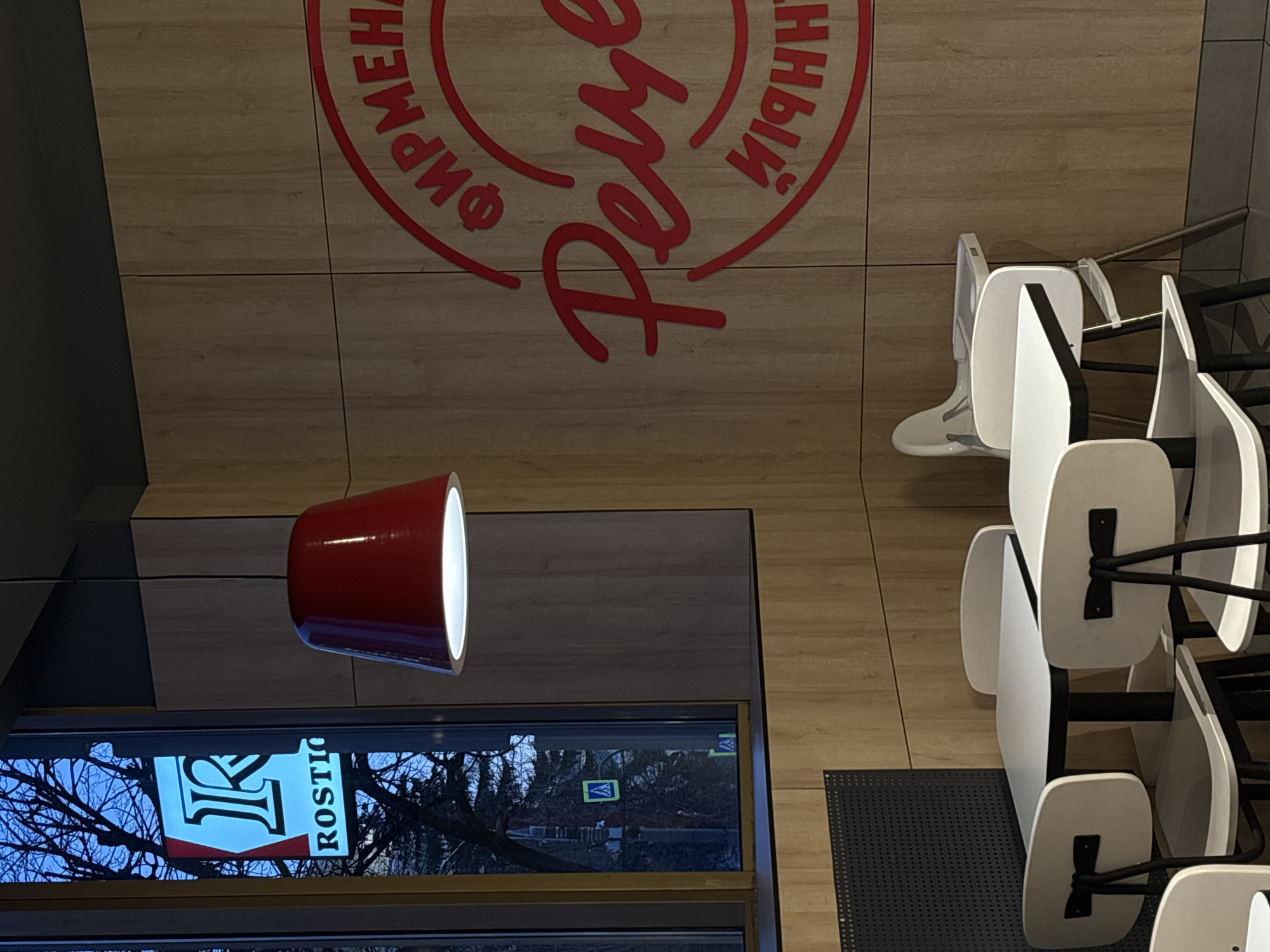 The image captures the interior of a fast-food restaurant or cafe, likely belonging to the Rostic's chain, located in Izhevsk, Russia. The scene is shot from a slightly elevated perspective, looking down.
The main subject is the floor, which features light wooden paneling adorned with a large, red circular logo. The logo prominently displays the stylized Cyrillic word "Ростикс" (Rostiks) in a script font, encircled by the words "ФИРМЕННЫЙ" (FIRMENNYY - "SIGNATURE" or "BRANDED") and "КУРИНЫЙ" (KURINYI - "CHICKEN"). This phrase translates to "SIGNATURE CHICKEN Rostiks."
To the right, several white chairs with black metal frames are visible, some appearing stacked or neatly arranged, suggesting the establishment might be closed, preparing to open, or undergoing cleaning. No people are present, indicating a quiet or off-peak time.
A prominent red, inverted conical pendant light fixture hangs from the ceiling on the left side of the frame, casting a bright light downwards. Below the light fixture, a large window offers a view to the outside. Through the window, bare tree branches are visible against a blue sky, suggesting either late autumn, winter, or early spring. A street sign, a blue square with a white pedestrian icon, is visible in the distance, indicating a pedestrian crossing. Reflected in the window is a vertical outdoor sign, partially visible, which clearly shows a large 'R' and the text "ROSTIC," further confirming the restaurant's branding.
The overall setting is clean and modern, with functional seating. The lighting through the window combined with the artificial light suggests it is likely evening or dusk.