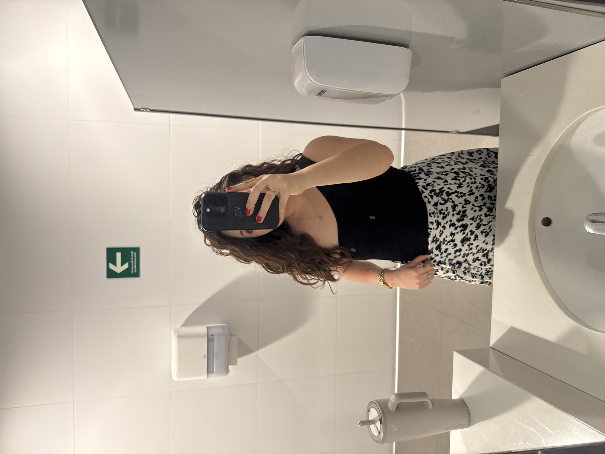 A woman is taking a mirror selfie in a bathroom. She is wearing a black top and a white and black skirt. The bathroom has white tiled walls, a sink, a hand dryer, a paper towel dispenser, and a green sign with a white arrow. The location is Mexico City, Mexico.