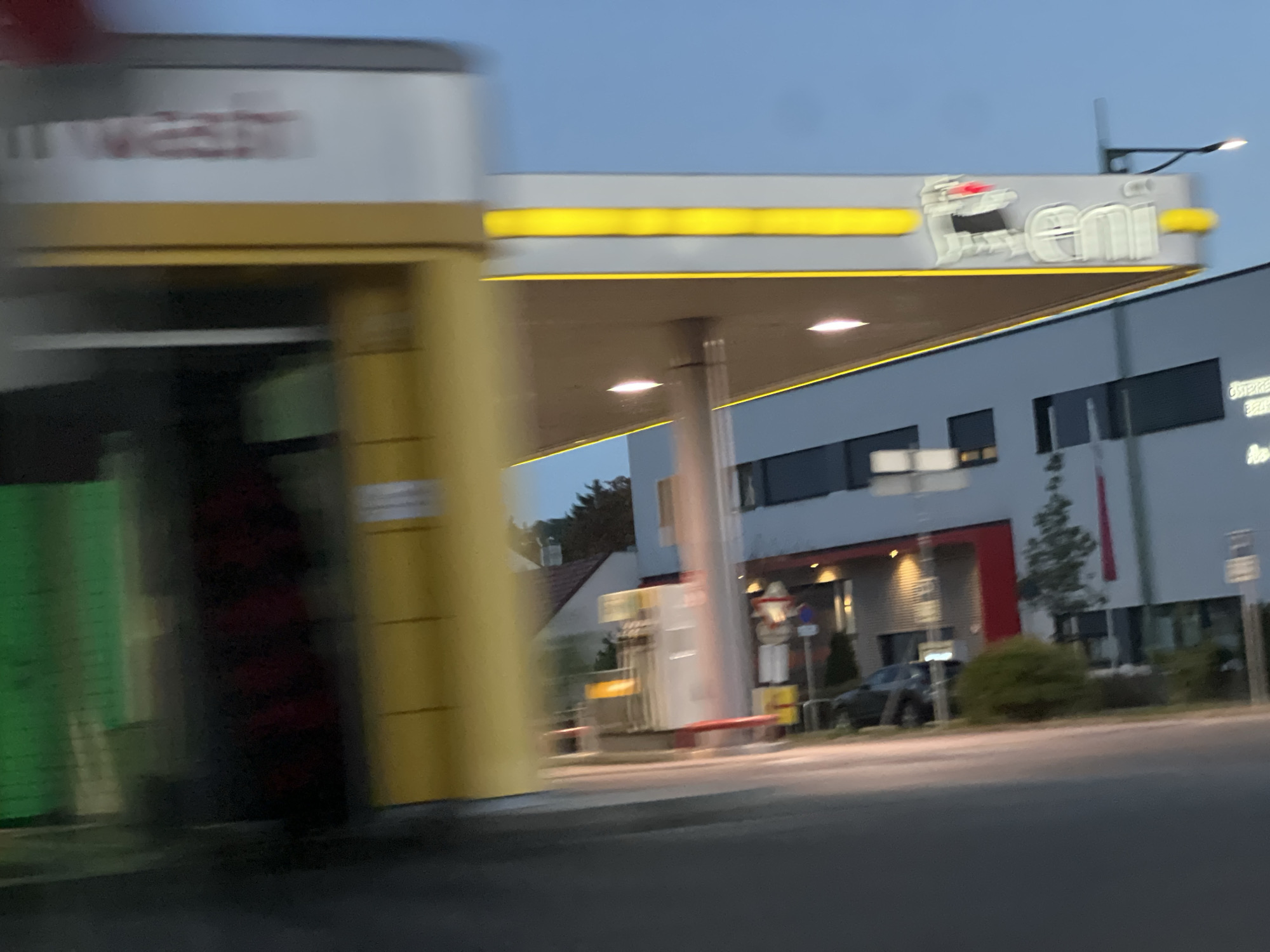 The image captures a blurred view of a modern gas station and car wash in Mödling, Austria. The prominent signage features a yellow horizontal stripe above a white panel with a red and white logo that appears to say "eni". To the left, a car wash structure with yellow and white elements is visible, with green and red brushes or mats partially obscured by motion blur. The background shows a building with dark windows and a red accent, a parked dark-colored car, and some greenery. The sky is overcast, indicating it is either dawn or dusk. The scene is captured from a moving vehicle, resulting in significant motion blur throughout the image.