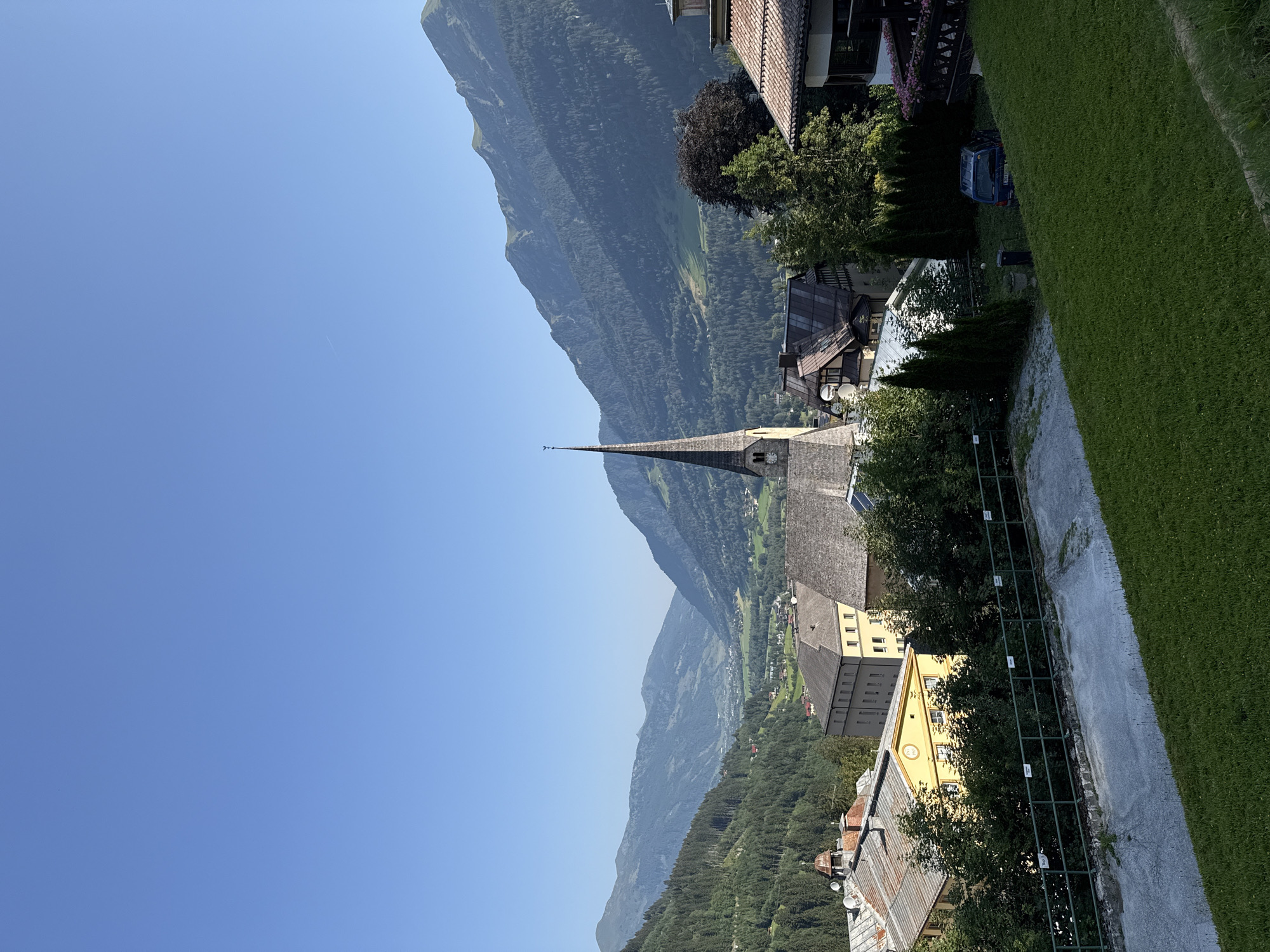 The image depicts a view of the town of Bad Gastein, Austria, on a clear day. In the foreground, a grassy slope leads down to a paved area and a green metal fence. Beyond the fence, several buildings are visible, including a prominent church with a tall, pointed steeple and a clock face. The church is situated in the center of the frame, surrounded by other structures with tiled roofs.
Further in the background, a picturesque valley opens up, framed by lush green mountains that are densely covered with trees. The mountains recede into the distance, with layers of them visible under a clear blue sky. The architectural style of the buildings suggests a traditional European alpine town. A dark blue van is parked to the right of the frame, partially obscured by trees. The scene conveys a sense of tranquility and natural beauty.