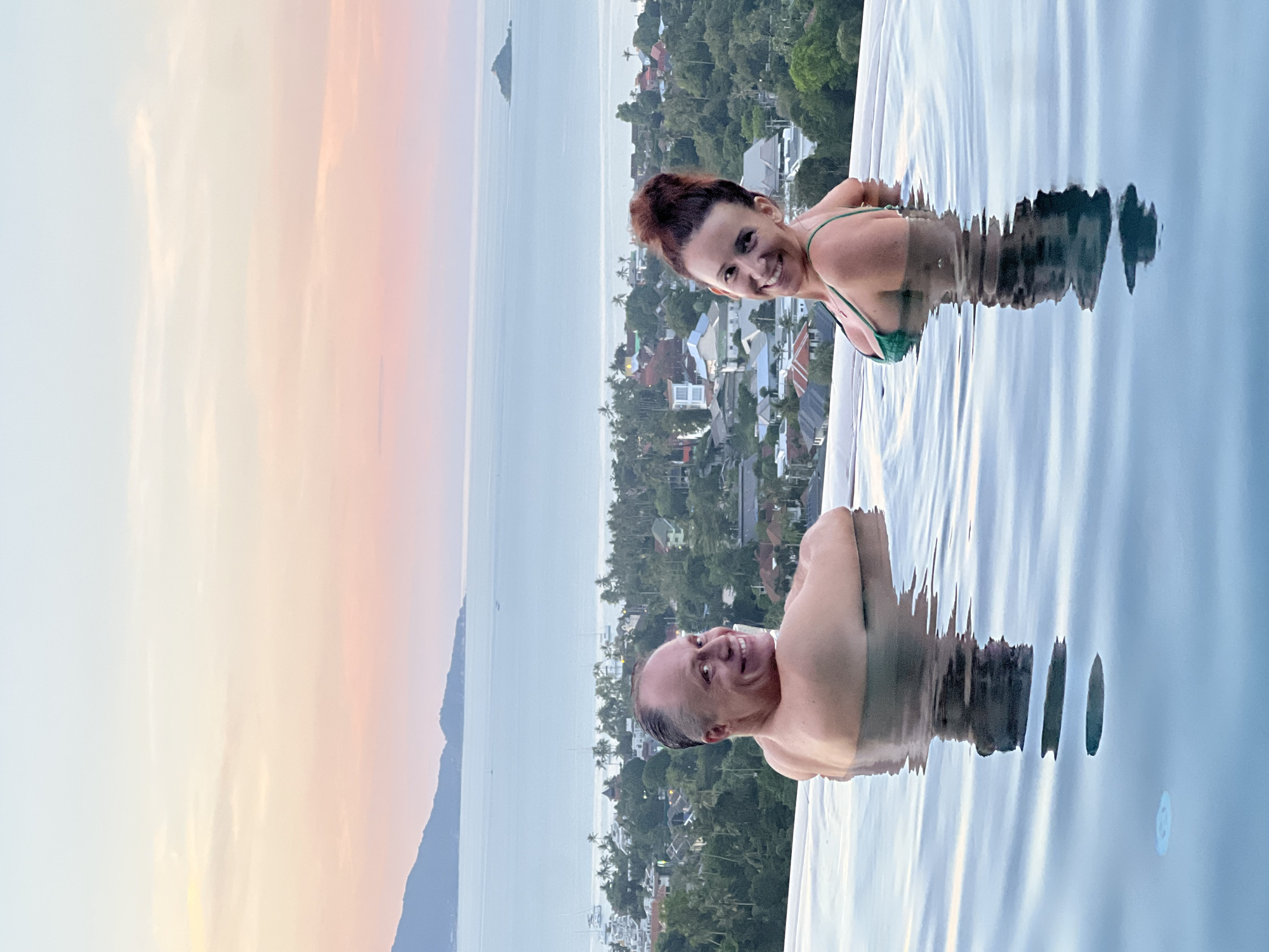 The image captures a man and a woman submerged in an infinity pool, with the ocean and a densely vegetated cityscape of Baan Bo Phut, Thailand, stretching out in the background. The sky displays a soft pastel gradient of pink, orange, and blue, indicative of either sunrise or sunset.
The man, positioned on the left, is looking over his left shoulder towards the camera with a wide smile. He is shirtless, and his upper body and head are visible above the water. The woman, on the right, is also smiling and looking towards the camera. She is wearing a green bikini top, and her upper body and head are visible. Both individuals are partially submerged in the clear blue water of the pool, which reflects the ambient light and the sky.
The cityscape below the pool is characterized by numerous buildings nestled amongst lush green trees and palm trees, suggesting a tropical environment. The ocean extends beyond the city, with several boats and islands visible in the distance. The water in the foreground of the pool shows ripples and reflections, creating a dynamic and serene atmosphere. The overall scene conveys a sense of relaxation and enjoyment in a picturesque tropical setting.