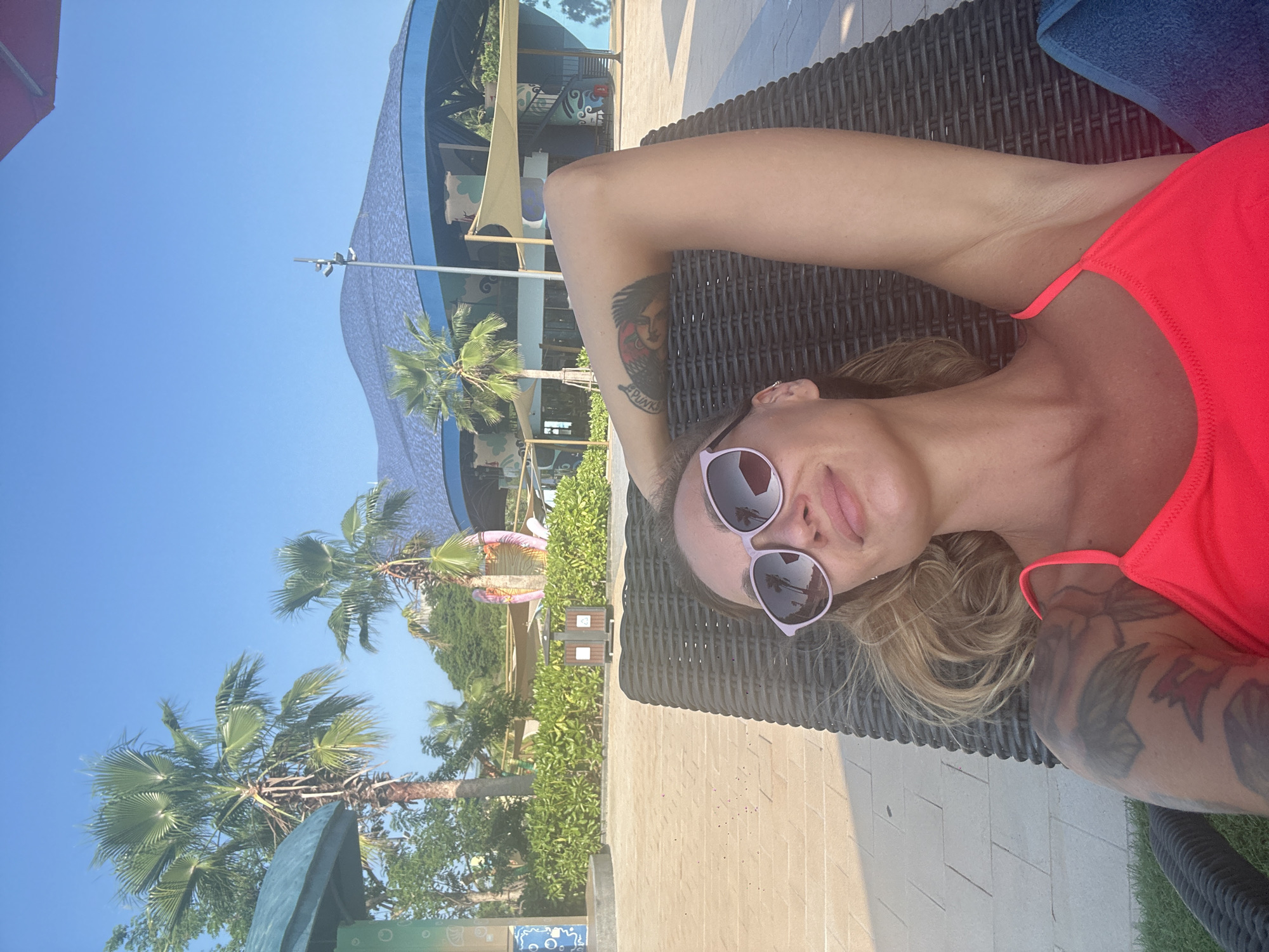 The image displays a female subject reclining on a dark brown woven lounge chair outdoors under a clear blue sky in Tien Thanh Ward, Vietnam. The woman has light-colored hair and wears purple-framed sunglasses with dark, reflective lenses, along with a bright neon pink/orange swimsuit top. Her left arm is bent, with her hand positioned behind her head, revealing a large dark tattoo on her upper arm and shoulder, which includes text partially discernible as "PUNK" and stylized imagery. To her right (viewer's left), a blue towel is visible on the lounge chair. The ground beneath and around the chair is covered with light-colored rectangular paver stones. In the background, numerous palm trees and green hedges are present. A dark brown waste bin stands on the paved area behind the woman. The scene also features a distinctive building with a large, undulating dark blue roof and light blue trim, featuring cream-colored fabric awnings and a visible dark blue spiral staircase on its right side. A colorful, abstract sculpture is partially visible among the trees further back. The environment suggests a resort or leisure area, well-lit by direct sunlight.