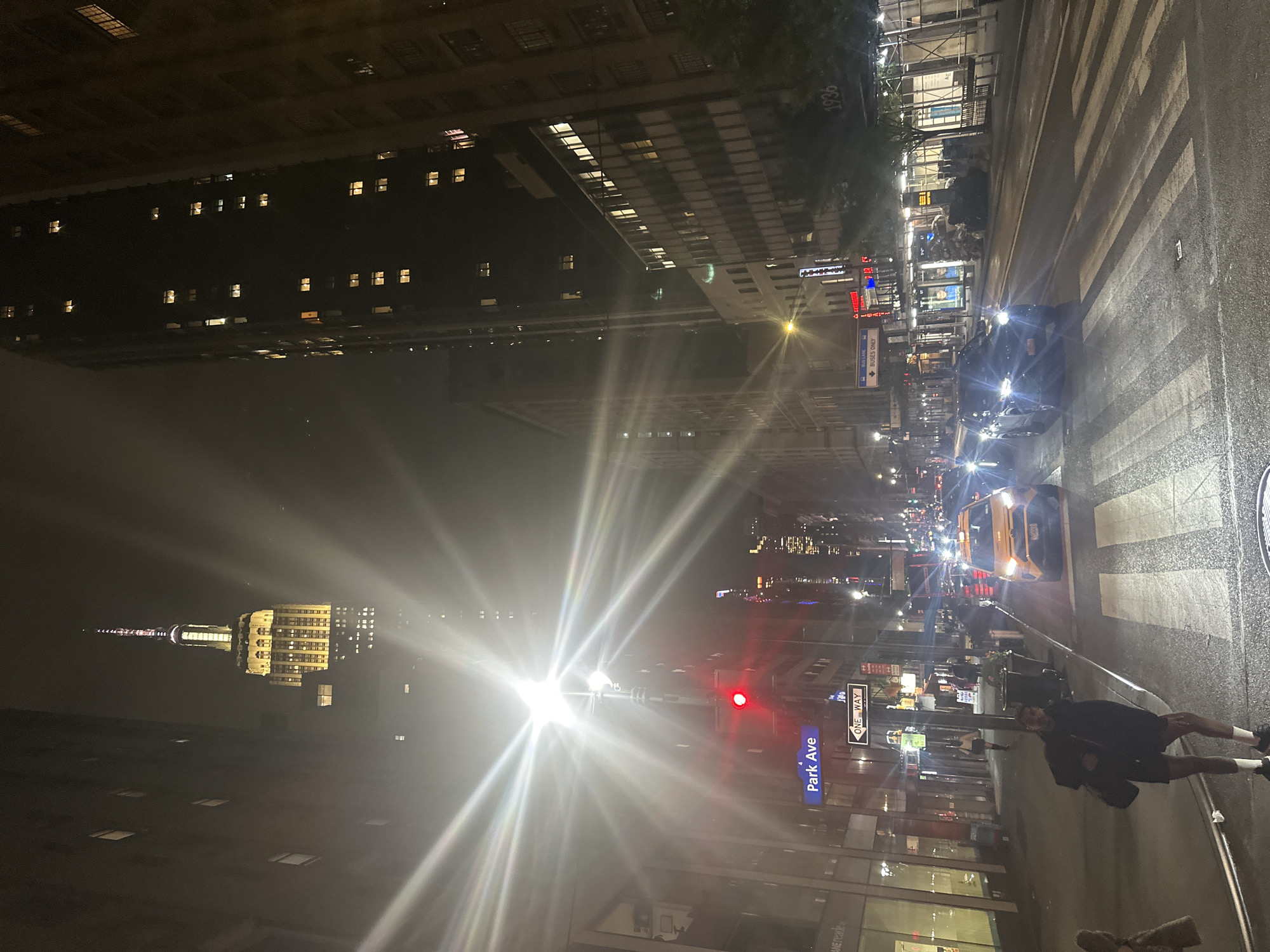 The image captures a vibrant night scene on a city street, likely in New York City, looking south on Park Avenue. The street is wet, suggesting recent rain or dampness. Tall, dark buildings with numerous illuminated windows line both sides of the avenue, while the iconic, brightly lit Empire State Building stands prominently in the distant left background.
In the foreground, a person is visible on the sidewalk to the right, dressed in dark shorts and a top, standing near the curb. Vehicles, including a distinctive yellow taxi cab, are present on the street, their headlights creating strong lens flares. A black car is closer in the mid-ground. Pedestrian crosswalks are marked on the asphalt.
Streetlights cast bright beams, and a red traffic light is visible on the right side of the street, indicating that traffic may be stopped or slowing down. Other distant lights from buildings and vehicles pepper the horizon, suggesting a bustling urban environment.
Visible text includes:
- A blue street sign reading "Park Ave".
- A white street sign indicating "ONE WAY".
- A blue sign with "BUSES ONLY" near the curb.
- The year "1936" is inscribed on the facade of a building.
The overall impression is a typical, dynamic city night, characterized by artificial lights reflecting off wet surfaces and the constant presence of traffic and pedestrians against a backdrop of towering architecture.