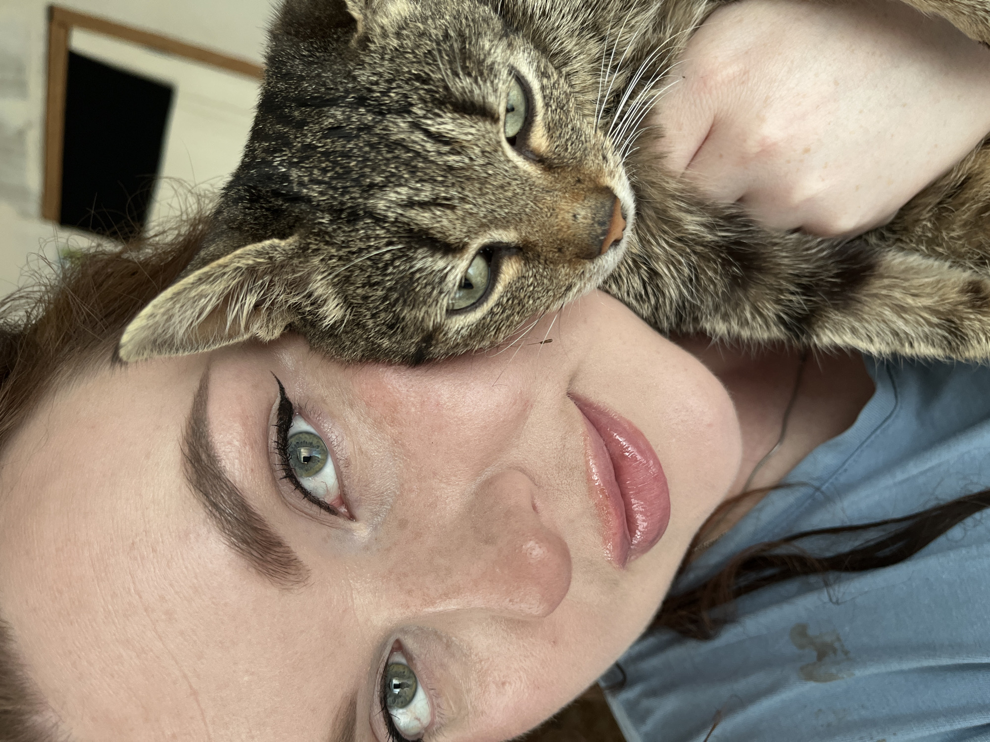 A light-skinned woman with reddish-brown hair holds a tabby cat close to her face. The woman has green eyes, winged eyeliner, and freckles. Her lips are glossy. The cat has green eyes and brown and black fur. The background is blurry and includes a picture frame on a wall and a light blue shirt. The scene is close-up, focusing on the faces of the woman and the cat. There is no visual information in the image to support the inclusion of Павловка, Russia, in the description.
