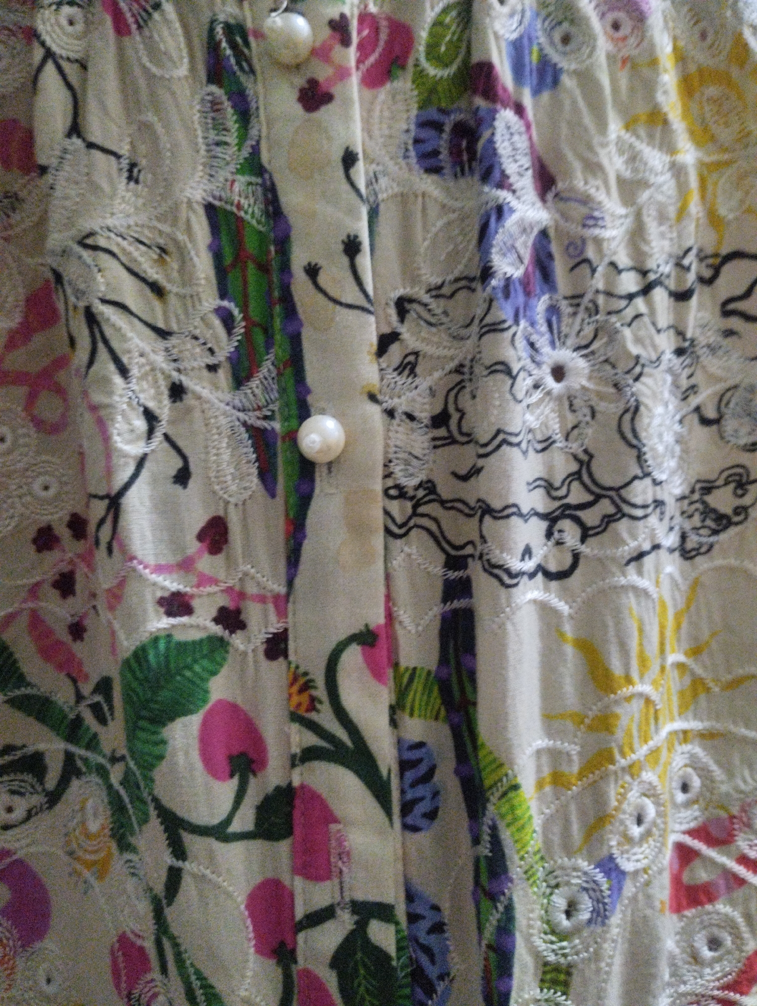 This media file is a close-up, vertical shot of a fabric with a colorful, abstract floral and botanical pattern. The fabric appears to be a light, possibly cream or beige, material. It is adorned with various embroidered elements and printed designs.
The pattern features a mix of solid color shapes and detailed embroidery. Prominent colors include vibrant pink, green, yellow, blue, and purple. There are stylized floral shapes in pink and yellow, as well as intricate green leaves and stems. Some areas have delicate white embroidery that outlines shapes or creates swirling patterns. Dark lines are also used in some embroidered sections, creating a graphic, almost calligraphic effect.
Two pearlized buttons are visible, positioned vertically down the center of the image, suggesting this fabric might be part of a garment, such as a shirt or dress. The lighting is soft and diffused, and there are no discernible shadows that would indicate a specific time of day or a strong directional light source. The background is out of focus, emphasizing the texture and design of the fabric. No people, specific settings, or weather conditions are visible. There is no visible text in the image.