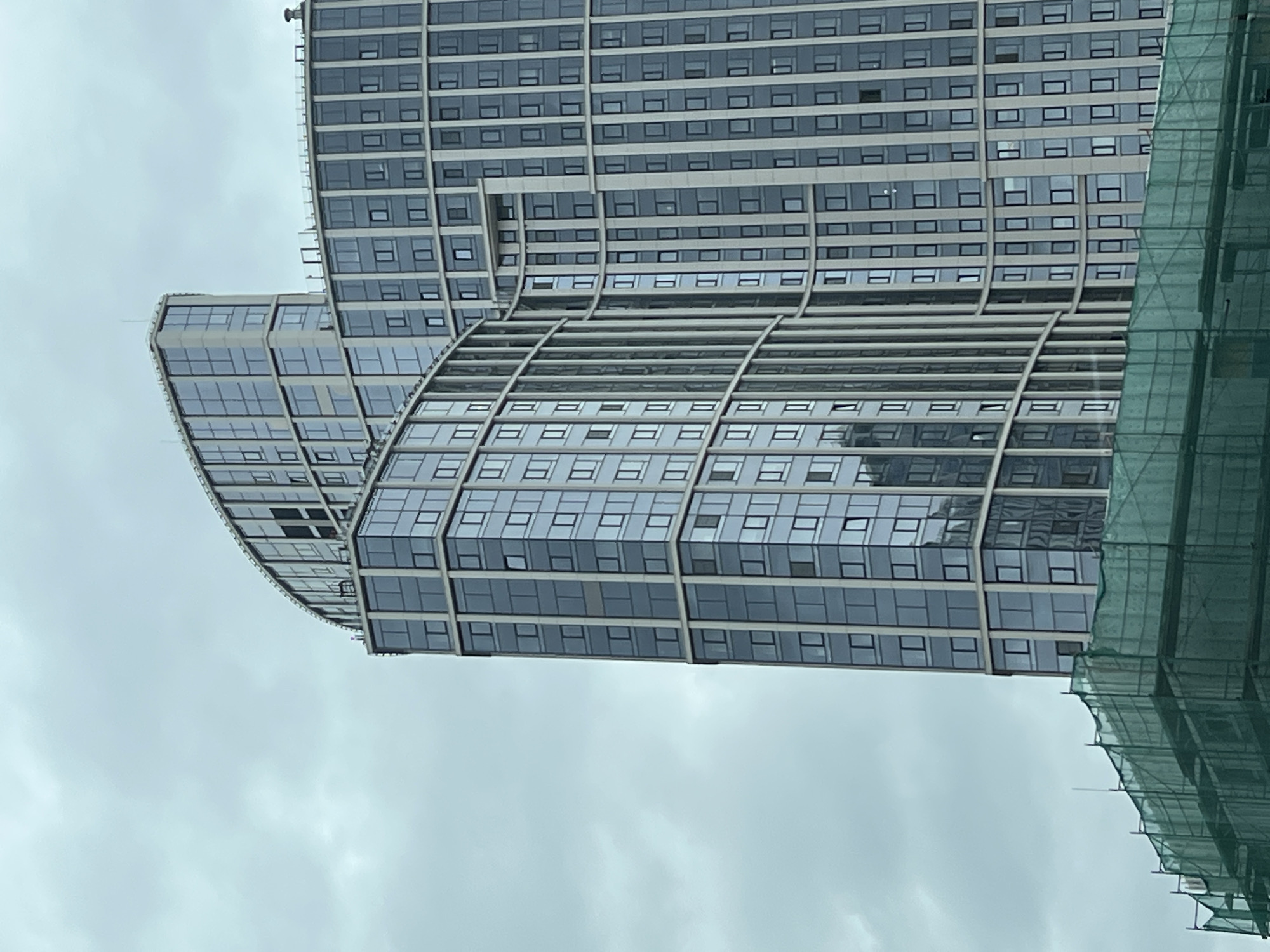 The image depicts a modern, high-rise building in Khimki, Russia. The architectural style is characterized by its curved, tiered structure with numerous rectangular windows. The facade is primarily glass and metal, giving it a contemporary appearance. Scaffolding with green netting is visible on one side, indicating ongoing construction or maintenance work. The sky is overcast, suggesting a cloudy day.