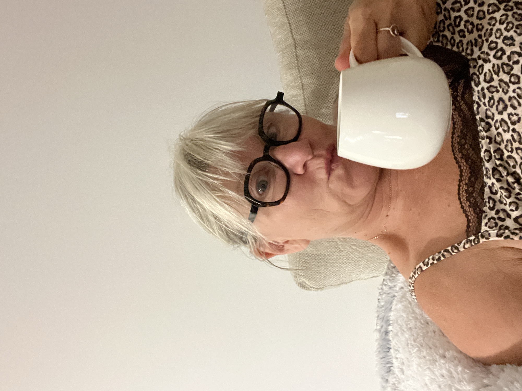 A woman with short, gray hair and black-rimmed glasses holds a white mug. She is wearing a leopard-print camisole with black lace trim. The woman is seated on a couch with a light-colored, textured cushion and a fluffy blue blanket. The background is a plain, off-white wall. The woman's facial expression is one of slight pursed lips. She is holding the mug with her right hand, and her left hand is not fully visible. The scene appears to be indoors, likely in a residential setting. There is no indication of the city of Cannes, France, in the image.