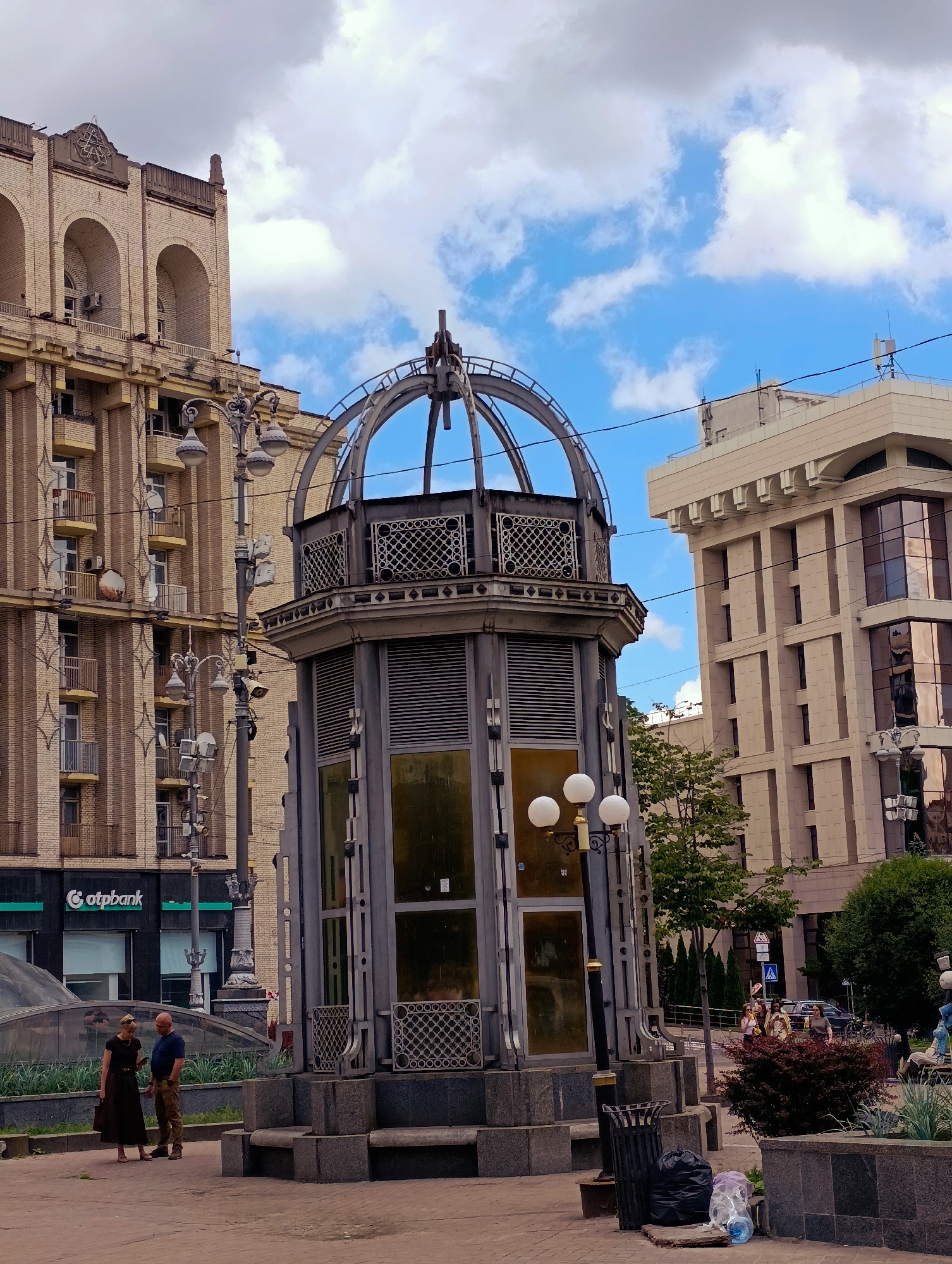 A couple stands on a paved plaza in front of a grey, octagonal structure with a domed roof. The structure has large, golden-tinted windows and decorative metalwork. Behind the structure, a tall, light-colored building with many windows and vertical decorative elements rises against a sky with white clouds. To the left of this building, a tan brick building with arched windows and balconies is visible. A street lamp with multiple globes stands to the left of the octagonal structure. In the foreground, there are planters with greenery, and a trash can overflowing with bags. The scene appears to be a public space in a city. The "otpbank" sign on the left building indicates the location is in Kyiv, Ukraine.