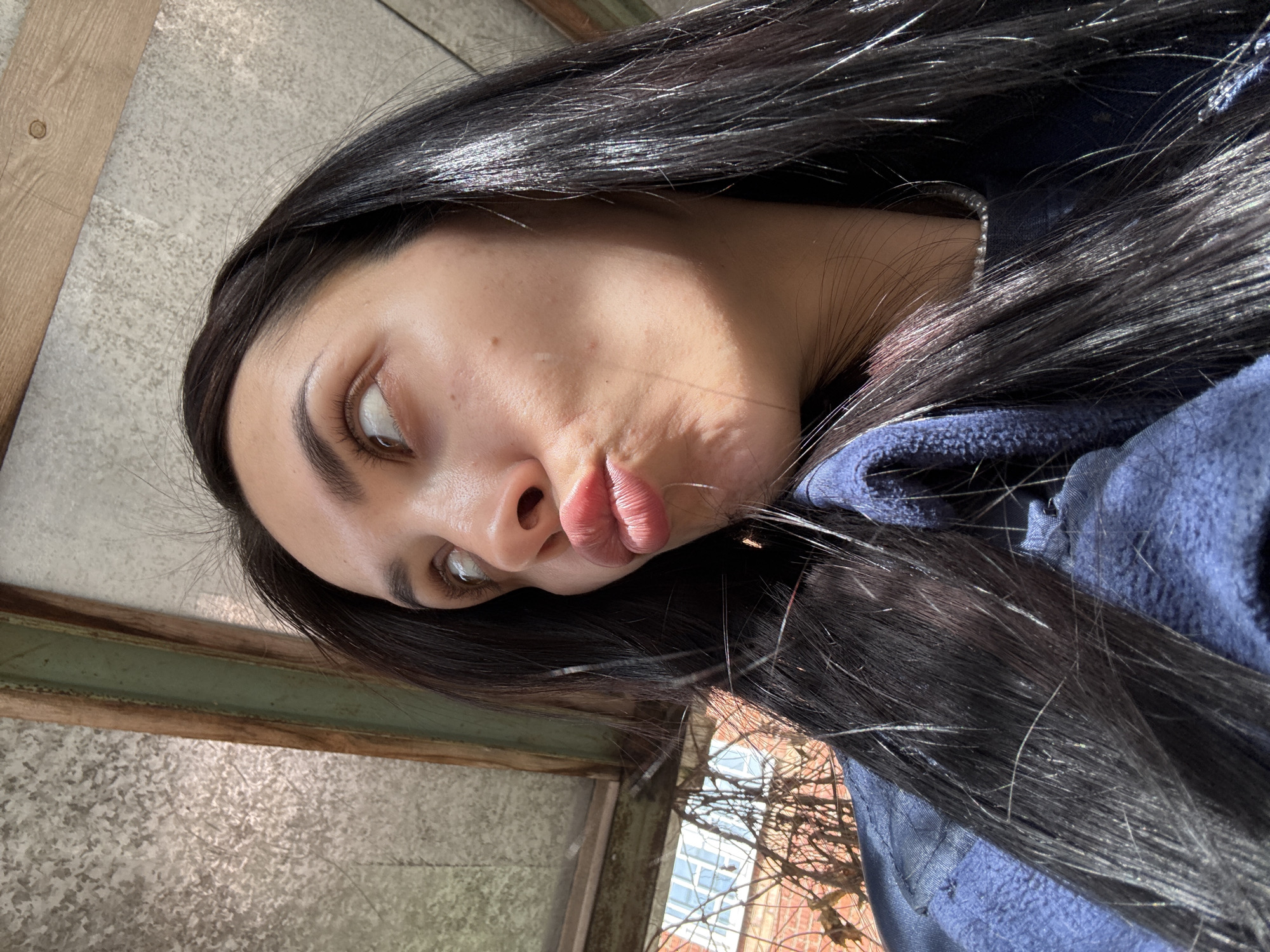 The image is a tilted portrait of a young woman with long, straight dark hair, captured from a low angle. She is looking upwards and slightly to her right, making a pouting expression with puckered lips. Her light brown eyes are visible, along with her neatly shaped dark eyebrows. Direct sunlight illuminates her face and hair, creating highlights on her dark, shiny hair and casting shadows that emphasize her facial features.
She is wearing a blue hooded garment, likely a fleece jacket or top, which is partially visible around her shoulders and intertwined with her hair. The background features various structural elements: a wooden beam or frame is visible in the upper left, set against a grey, textured surface, possibly concrete or stone. Another greenish-brown beam runs horizontally across the middle left of the frame. Beyond these structures, a view through what appears to be a window or opening reveals parts of a red brick building and numerous bare tree branches against a bright sky. The setting suggests an outdoor or semi-outdoor location, possibly a balcony, shed, or courtyard. No visible text is present. The time of day appears to be sunny daytime, and the location is Votkinsk, Russia.