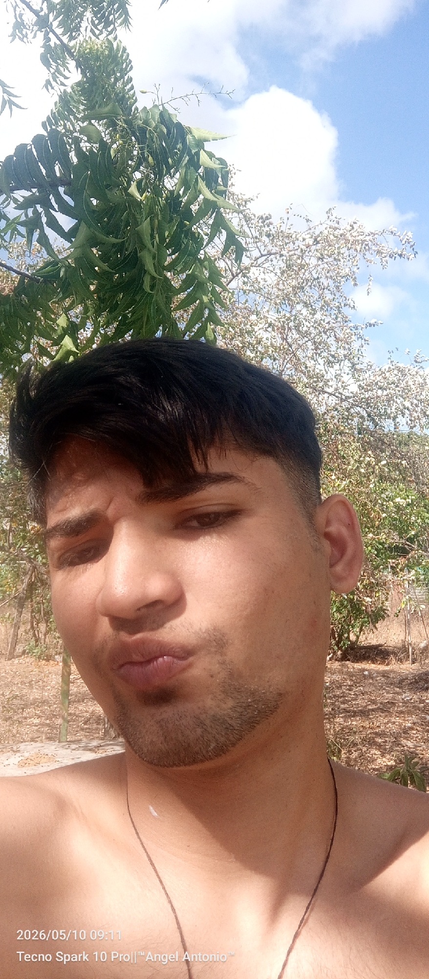A man is posing outdoors for a selfie-style photo. He has dark hair, light skin, and a short beard. He is shirtless and wears a thin necklace. His lips are pursed. Behind the man are trees, one with green leaves and another with white flowers. The sky is blue with scattered white clouds. The bottom of the image contains a timestamp "2026/05/10 09:11" and device information "Tecno Spark 10 Pro|| "Angel Antonio"". Juan Griego, Venezuela is inferred from the timestamp and device information.