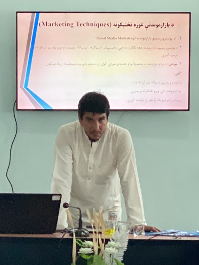 Here is a description of the image:
The image appears to be taken indoors, possibly in a classroom or conference room in Shurandam, Afghanistan, given the language on the projection screen. A middle-aged man with short dark hair and wearing a long-sleeved white shirt is standing behind a table or lectern. On the table, there is a laptop, a microphone, a vase of flowers, and a glass. Behind the man, there is a projection screen displaying text in what appears to be Pashto, and the English translation, "(Marketing Techniques)". The text on the screen seems to outline strategies for social media marketing. The lighting is somewhat subdued, and the walls are a light green color. The focus is on the man and the presentation screen.
