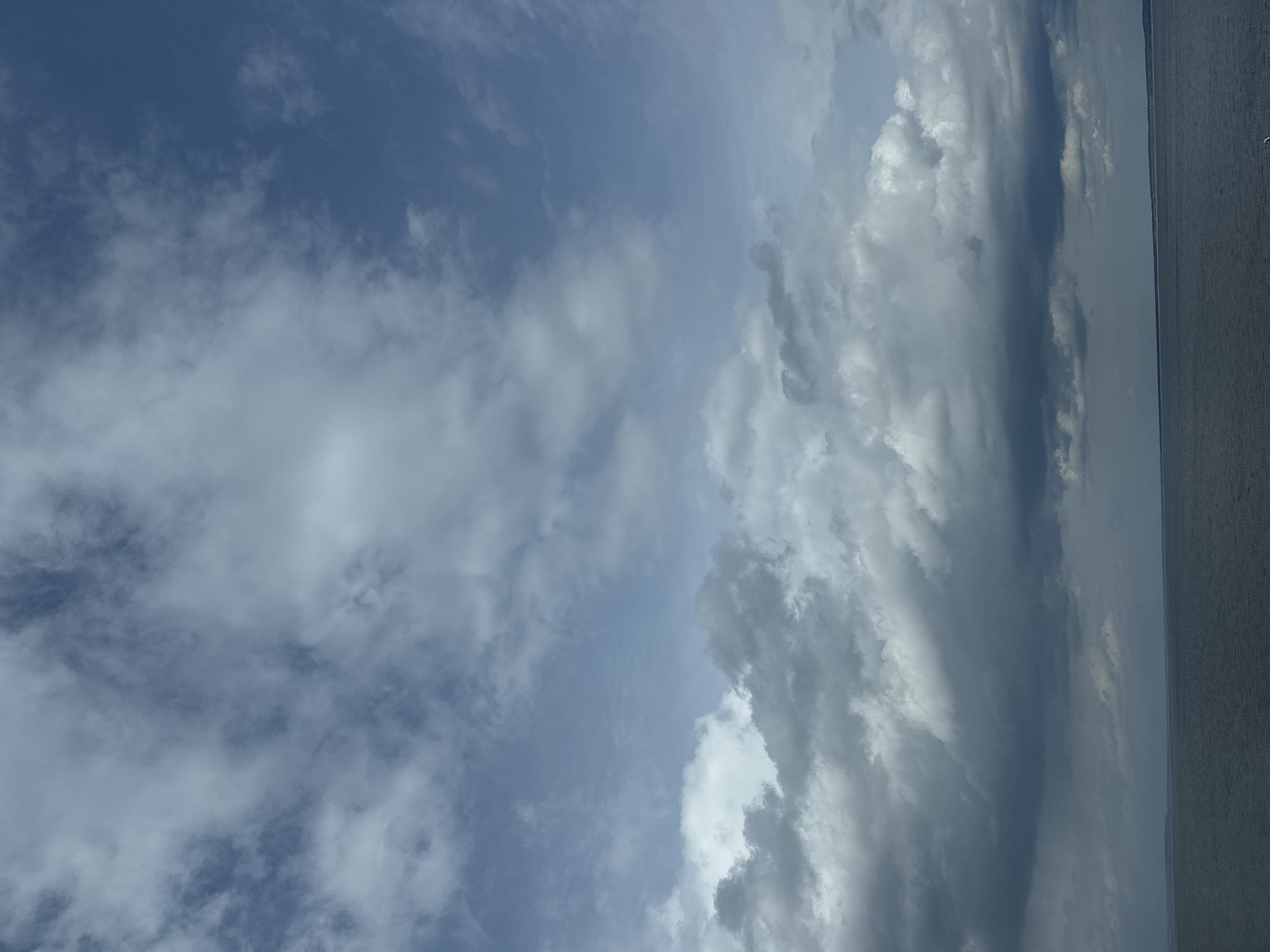 The image shows a cloudy sky over water. The left side of the image features patches of blue sky with wispy clouds. The right side is dominated by a bank of thick, grey storm clouds. The water is relatively calm. There are no people, subjects, objects, or actions. The location is implied to be in the city of Chaikovsky, Russia.
