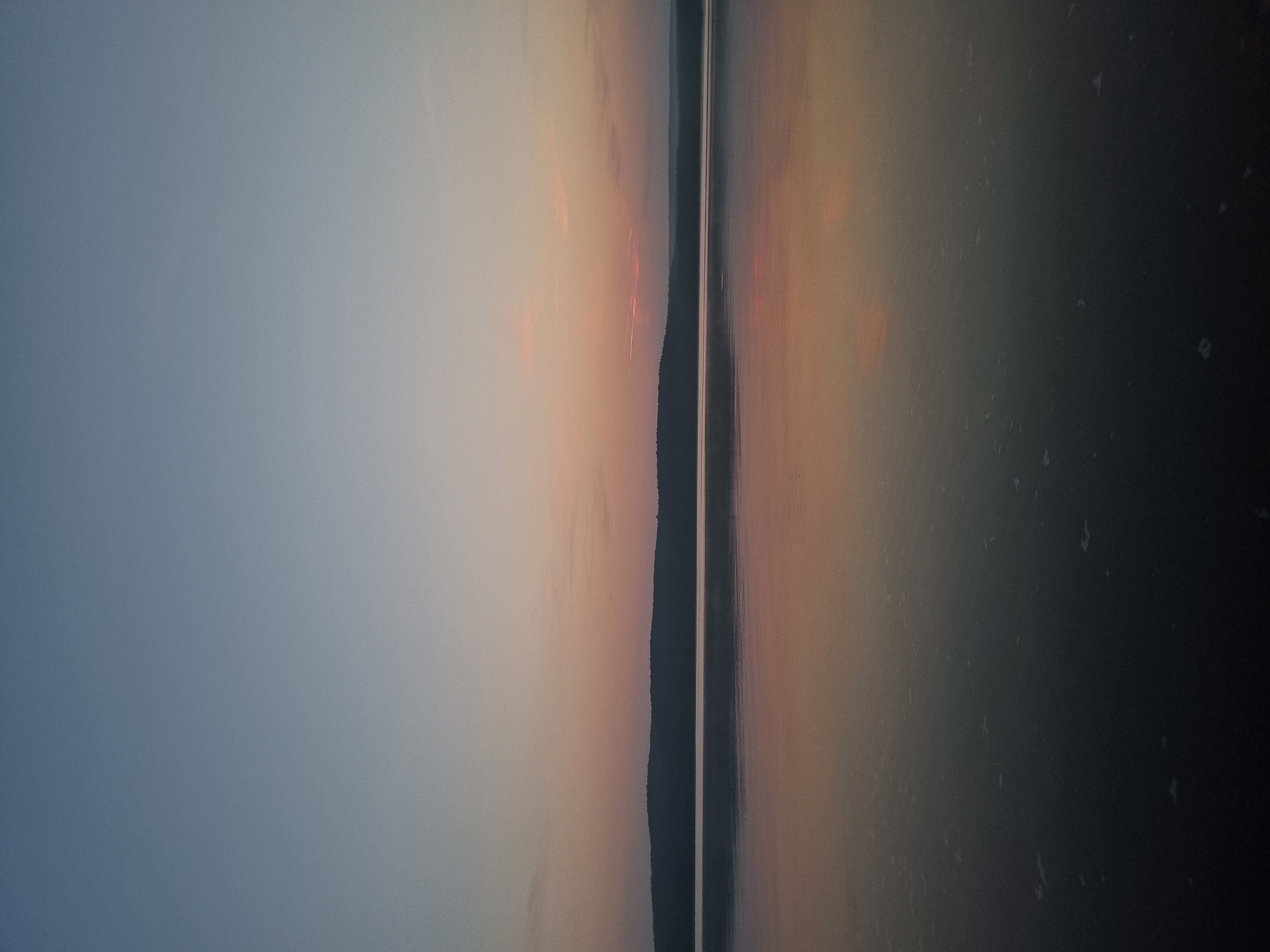 The image depicts a serene natural landscape at dusk, featuring a calm body of water reflecting the muted colors of the sky. A dark, treed silhouette of a distant shoreline forms the horizon line. The sky transitions from a pale blue at the top to soft hues of pink and orange closer to the horizon, suggesting either a sunrise or sunset. The water's surface is mostly smooth, with subtle ripples and some small, light-colored patches that could be foam or debris. The overall impression is one of tranquility and natural beauty, devoid of any human presence or structures. This scene is consistent with views along the Kama River in the city of Chaykovsky, Russia, known for its expansive water bodies and natural surroundings.