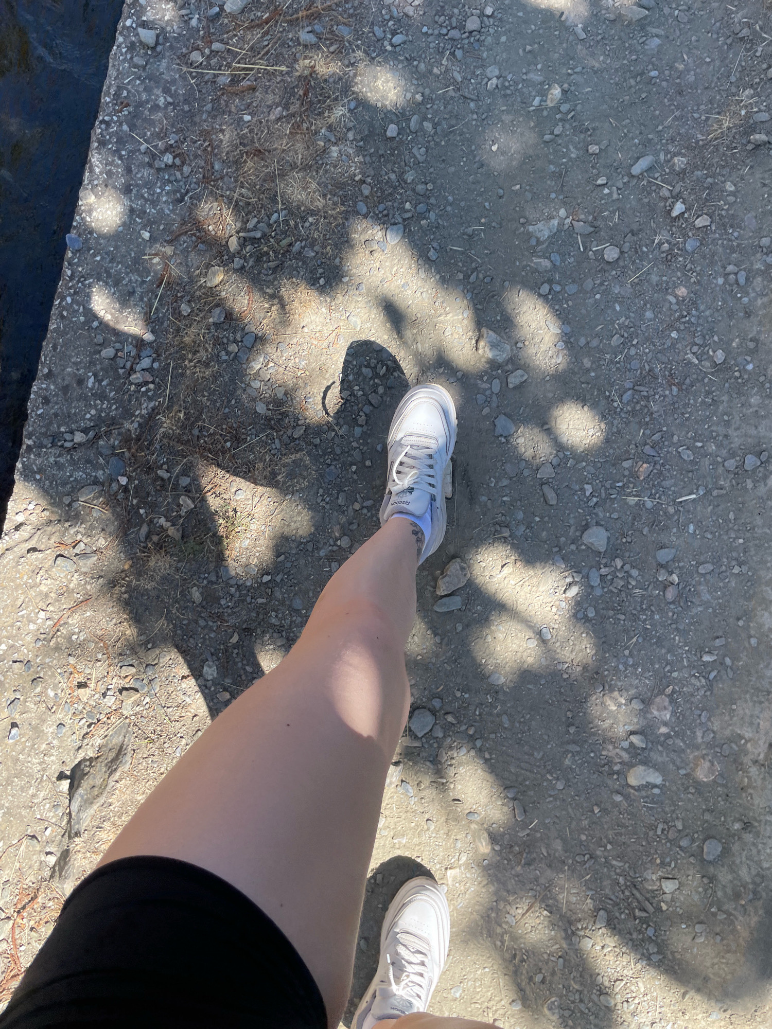 The image shows a person's legs and feet as they walk on a gravel path next to a concrete edge. The path is covered with small stones and dirt, with shadows from trees dappling the ground. The person is wearing black shorts and white athletic shoes. The concrete edge appears to be the border of a body of water, possibly a canal or stream. The scene suggests an outdoor activity like walking or hiking, possibly in a park or natural area. The provided metadata indicates the location is Lanjarón, Spain.