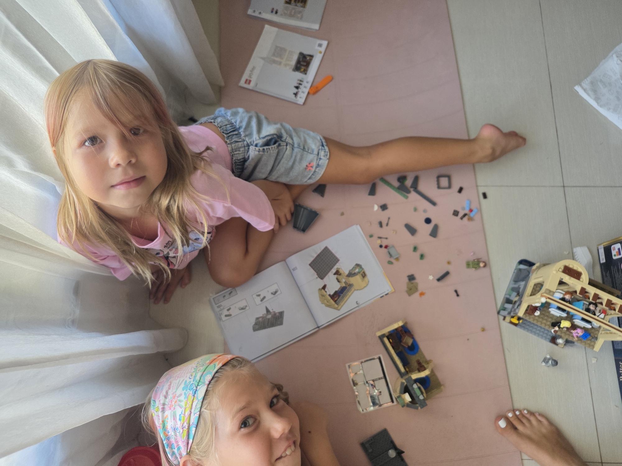 The image depicts two young girls on a pink rug in what appears to be a room in North Kuta, Indonesia. They are constructing a LEGO set, evidenced by the LEGO instructions and scattered pieces. One girl is looking at the camera, wearing a pink shirt and denim shorts; the other is wearing a patterned headband. A partially completed LEGO building lies on the floor, along with the LEGO box. The flooring transitions from the pink rug to ceramic tiles. Curtains are visible in the background.