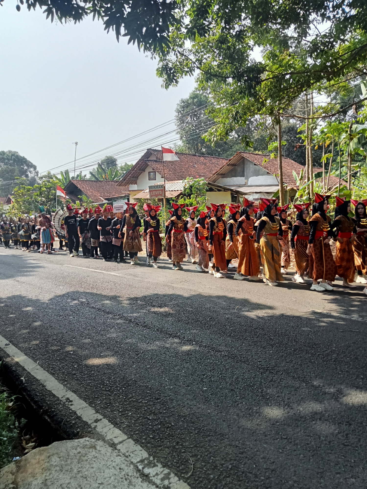 An outdoor, eye-level, vertical, medium shot captures a procession of people in traditional attire walking along a paved road in Dororejo, Indonesia. The procession features a group of women in the foreground, adorned in patterned orange and brown dresses and red headpieces. They are followed by men and children, some wearing black shirts and others in more elaborate costumes, including one individual with a large, circular headdress. A drum can be seen at the front of the procession.
The environment consists of a paved road with a white stripe marking the edge. On the right side of the road, houses with red tile roofs are visible, along with trees and lush green foliage. The sky is clear and bright, suggesting a sunny day. The scene appears to be a cultural parade or festival, likely celebrating a local event or holiday. The overall atmosphere is festive and communal.