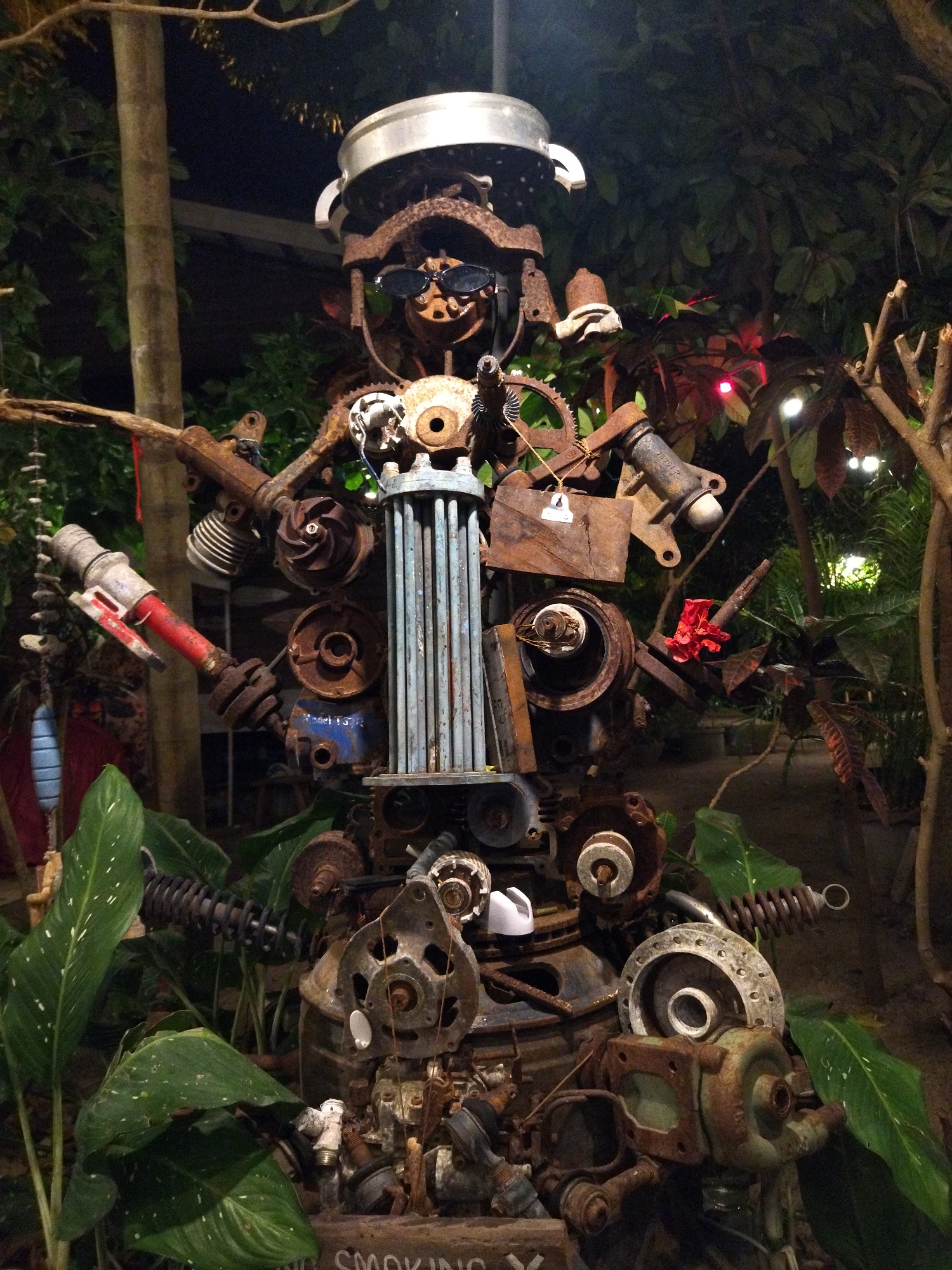 This photo captures a bizarre, life-sized sculpture crafted from rusty scrap metal, located in ตำบลเกาะพะงัน, Thailand. The sculpture features a variety of metal parts, including gears, pipes, springs, and wheels, assembled into a humanoid form. The sculpture is adorned with accessories such as sunglasses and a cooking pot on its head. The base of the sculpture is surrounded by green plants. At the base is also a sign that says “SMOKING X”. In the background, there are trees and foliage.