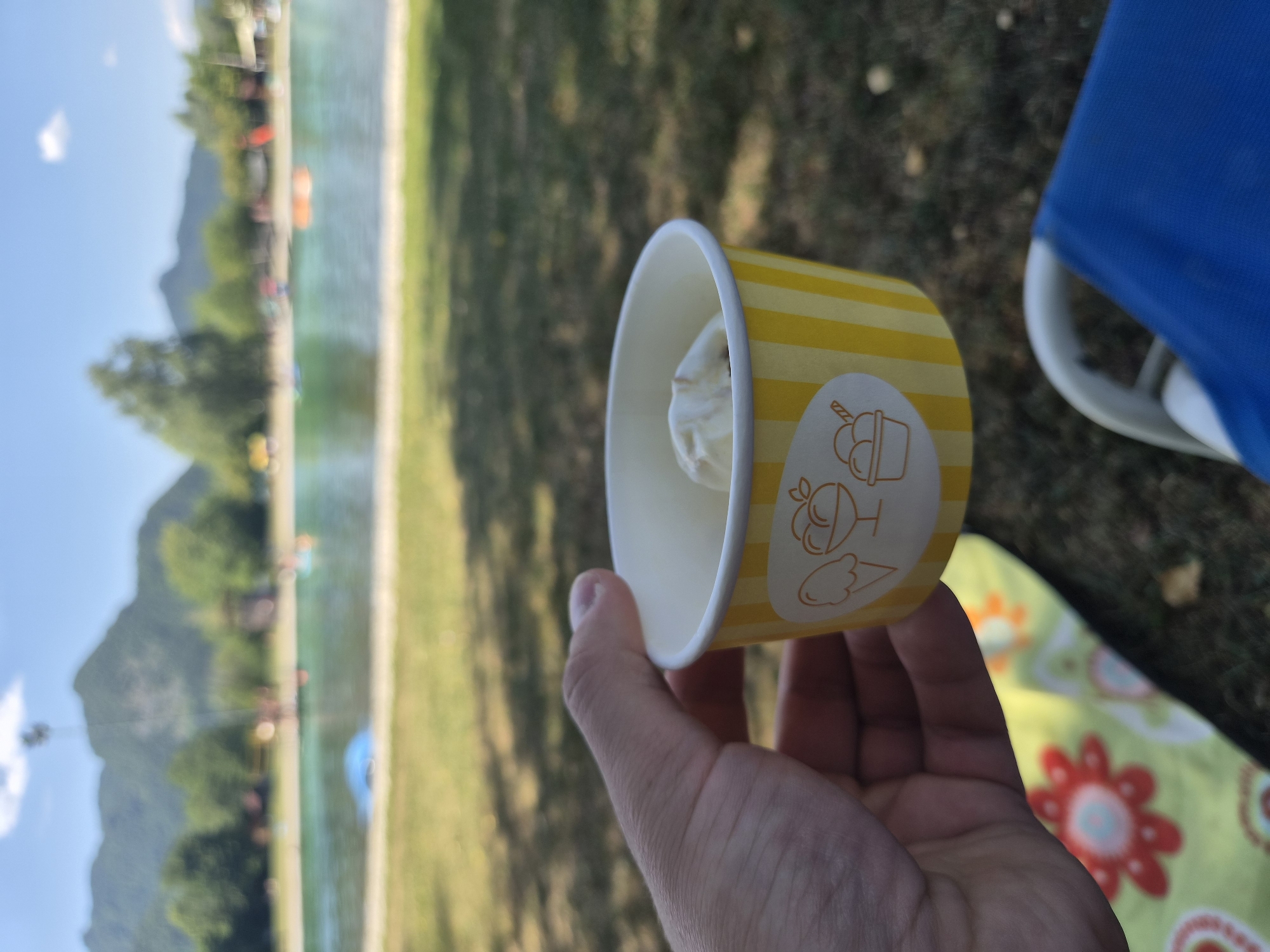 In Landl, Austria, a person is holding a yellow and white striped paper cup filled with ice cream. The cup has an illustration of ice cream cones and cups on it. In the background, a swimming pool or lake is visible with people swimming and enjoying themselves. Mountains and trees form the backdrop of the scene. A blue chair is partially visible in the foreground.