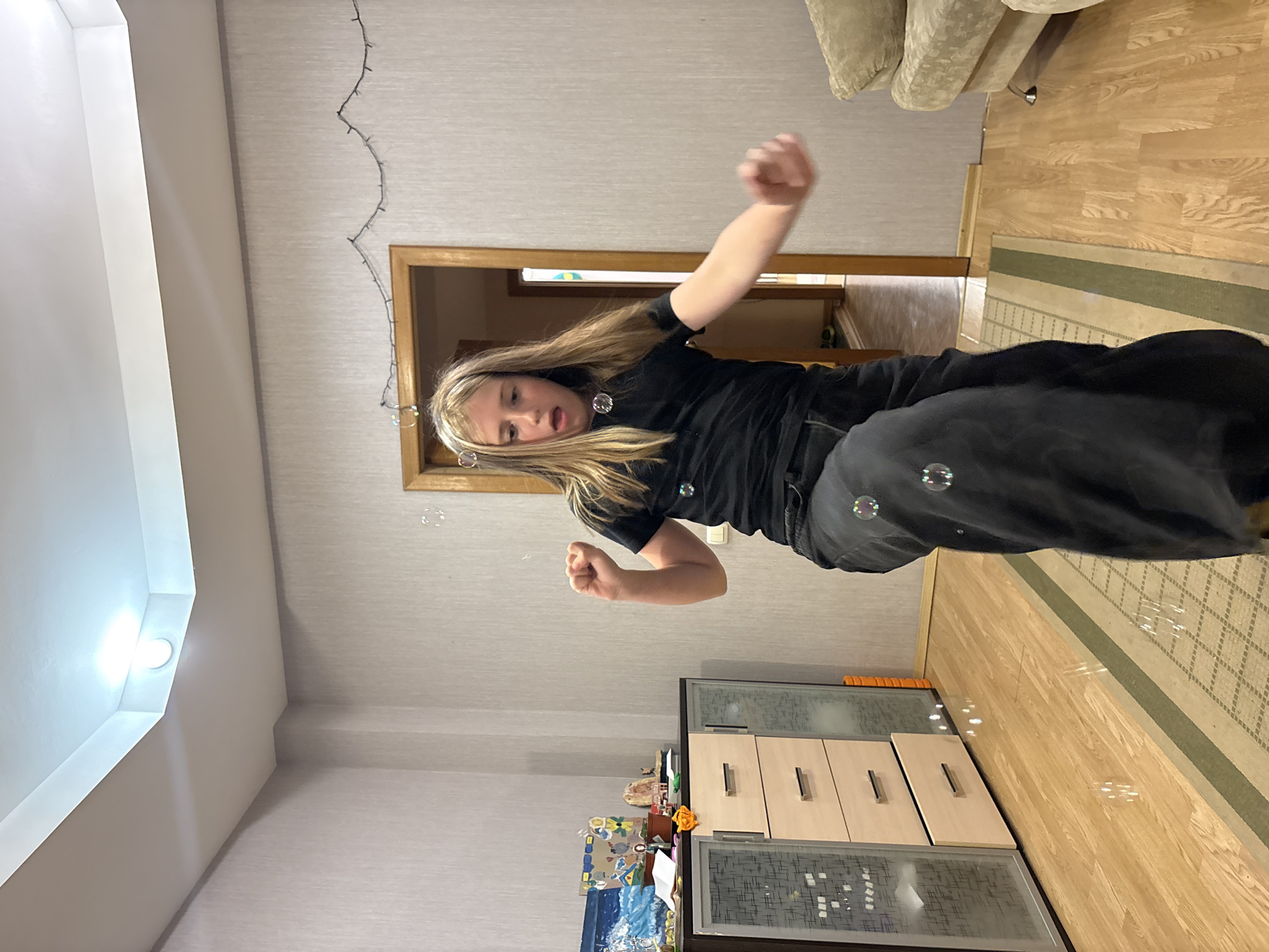 In a brightly lit room with beige textured wallpaper, a young person with long blonde hair, wearing a black t-shirt and dark pants, is in motion, possibly dancing or playing, with several soap bubbles visible around them. The person's mouth is open, suggesting exertion or surprise. The room contains a tall, dark wood cabinet with several drawers and glass-fronted sections on the left, a doorway leading to another room in the background, and a portion of a light-colored sofa on the right. The floor is polished wood, with a striped rug running through the center of the image. The ceiling features recessed lighting and a decorative trim. The overall impression is of a casual indoor activity. The location in Veliky Novgorod, Russia, cannot be determined from the visual information provided in the image.
