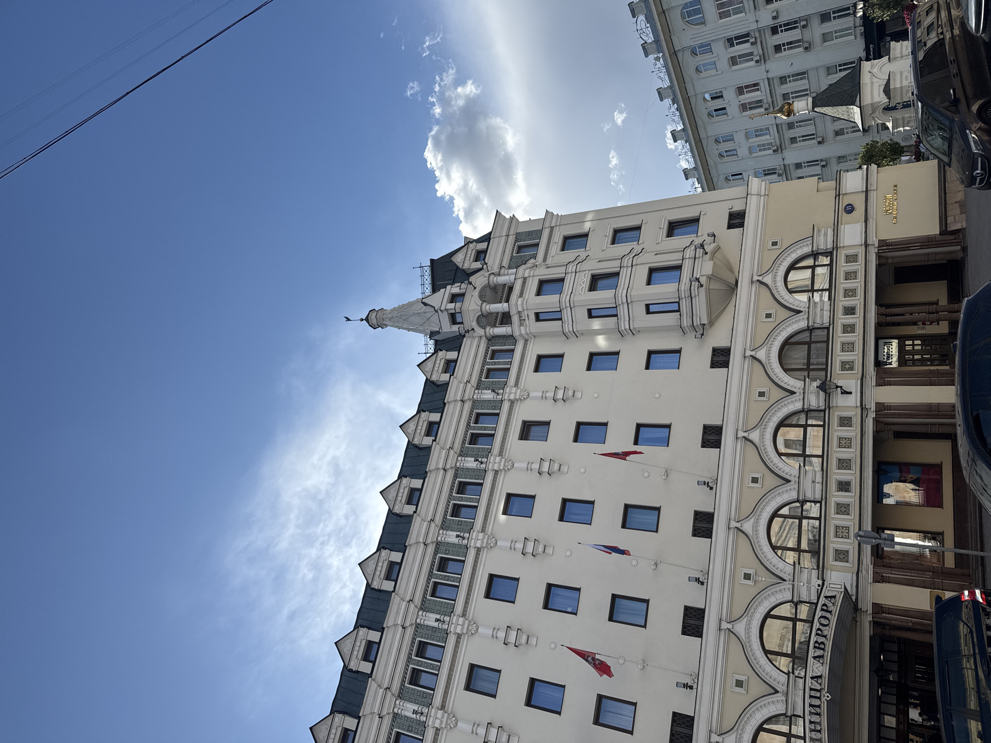 This photograph shows the exterior of the Hotel Aurora, located in Moscow, Russia. The building is tall, multi-storied, and light-colored, with a steeple-like architectural element extending from the top. Rows of windows, some with Russian flags hanging below, are visible on the facade. The lower section features arched windows and an entrance. The sign "Hotel Aurora" is displayed. The sky is visible above the building with a blue expanse and some clouds. Other buildings and vehicles are partially visible on the right side of the image. Powerlines are visible in the upper left corner.