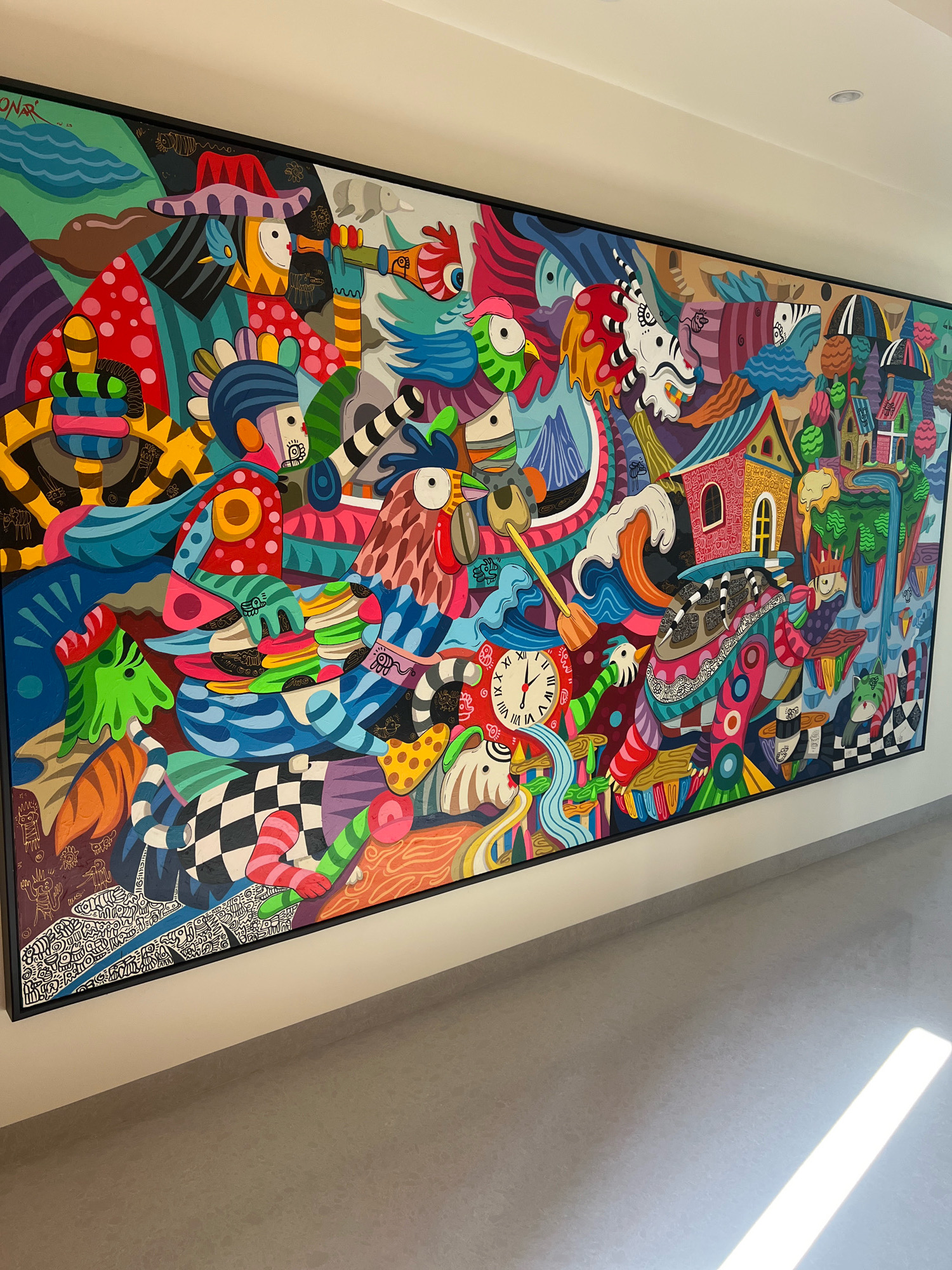 The image shows a large, vibrant, and intricately detailed mural painting with a whimsical and surreal style. The painting is mounted on a white wall in a hallway with a gray floor. Sunlight streams in from the right side of the frame, creating bright highlights on the floor.
The mural depicts a chaotic yet harmonious composition of various fantastical creatures, human-like figures, and architectural elements. There are several bird-like figures with large eyes, some with colorful plumage and beaks. One prominent figure wears a wide-brimmed hat and peers through a telescope. Another section features a clock with Roman numerals. There are also houses with pitched roofs, some with windows and doors, integrated into the fantastical landscape. Abstract shapes and patterns, such as checkered squares and stripes, are woven throughout the artwork. The color palette is rich and varied, with prominent use of blues, reds, yellows, greens, purples, and pinks. The painting is framed by a thick black border.
The context of the location in Ngaglik, Indonesia, is not visually present in the image itself. The provided description is based solely on the visual content of the photograph.