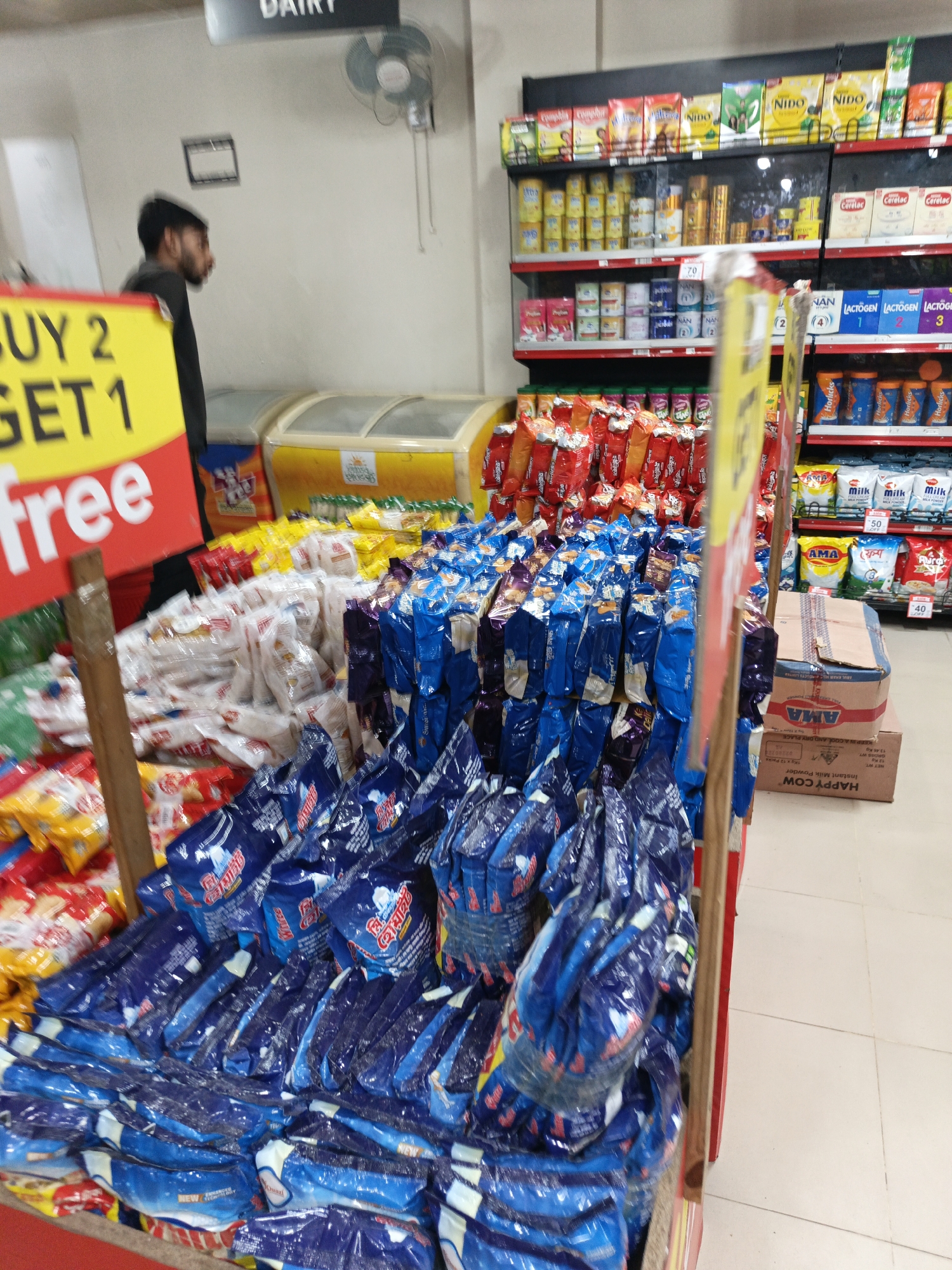 This is an indoor shot taken in what appears to be a grocery store or supermarket, likely in Dhaka, Bangladesh. The store features shelves stocked with various food products, including milk powders and infant formula. A promotion sign with the text "BUY 2 GET 1 free" is prominently displayed. The image shows a display of packets, with mostly blue packaging. A man in a black shirt is seen in the background near the dairy section, as indicated by the "DAIRY" sign. There are cold storage units with yellow tops next to the aisle. Boxes of AMA brand milk powder and other milk products are visible on shelves in the background.