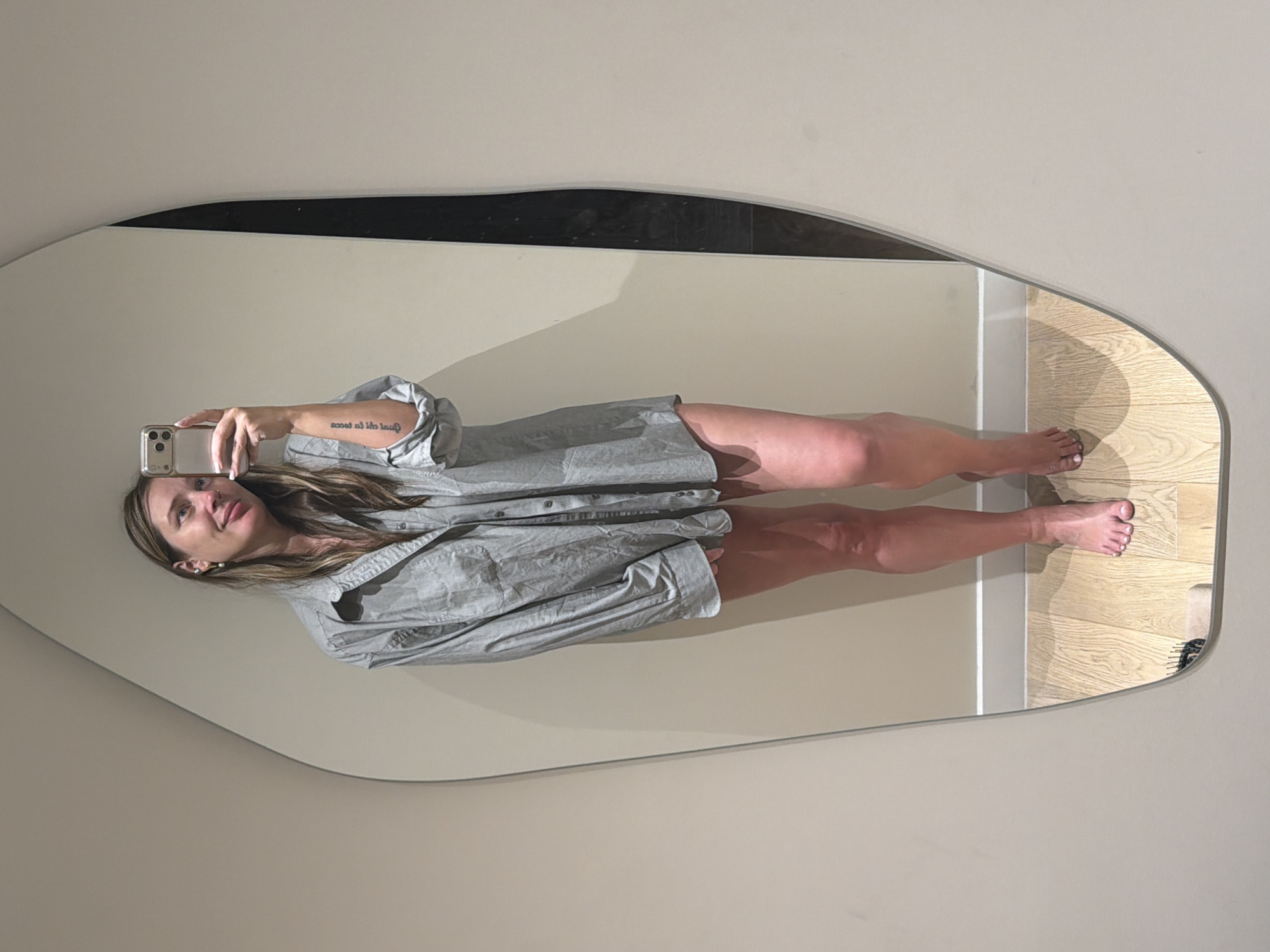 A woman is taking a mirror selfie in a room with beige walls and light wood floors. She is wearing an oversized light blue button-down shirt and is barefoot. She has long brown hair and is holding a silver smartphone in her right hand. A tattoo is visible on her left forearm. The mirror is irregularly shaped and mounted on the wall. The image does not contain any information that could indicate the location in Moscow, Russia.