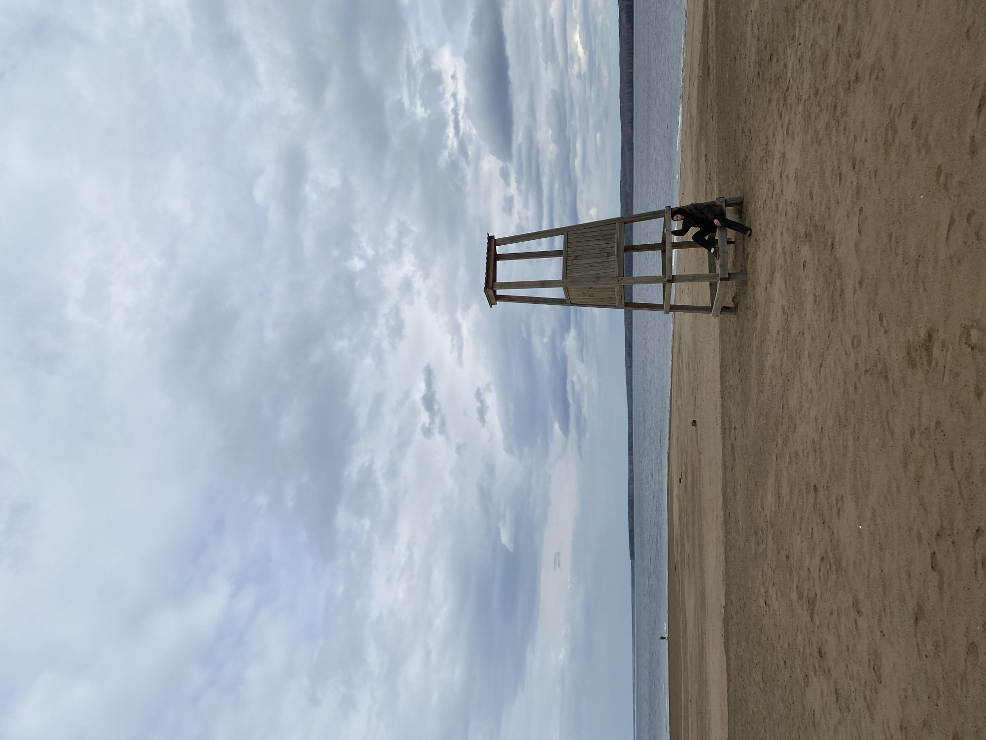 A wooden lifeguard tower stands on a sandy beach in Chaikovsky, Russia. A person in dark clothing and shoes climbs on the structure. The sky is cloudy, and the water is calm.