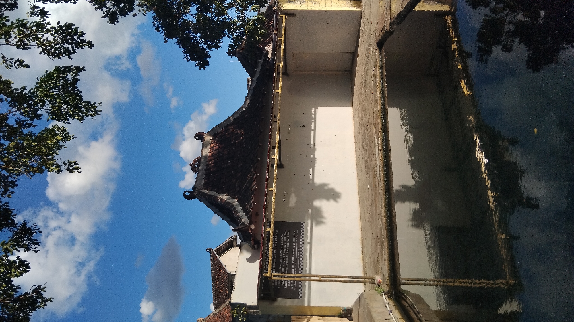 This is a vertical, eye-level, outdoor, medium shot photograph of a traditional building and a water feature. The building has a dark tiled roof with ornate, curved ends. The walls of the building are white, with a darker gray sign mounted on the left side. The sign has text in Indonesian, which appears to be rules or information about the area. A golden horizontal beam runs across the top of the white wall, supporting the roof overhang. To the right of the main building, a smaller, peaked roof of similar construction is visible.
In the foreground, a large, dark body of water reflects the building and the sky. The water is calm, with ripples creating distorted reflections of the surrounding structures and trees. The ground surrounding the water is paved with light-colored bricks.
Above the building, the sky is a vibrant blue with scattered white, puffy clouds. Branches of a leafy green tree extend into the frame from the top and right edges of the photograph, casting shadows on the building.
The scene is likely located in a rural or semi-rural area in Ngaglik, Indonesia, given the traditional architectural style and the presence of a water feature, possibly a spring or a communal bathing area. There are no people visible in the image. The overall impression is one of tranquility and cultural heritage.