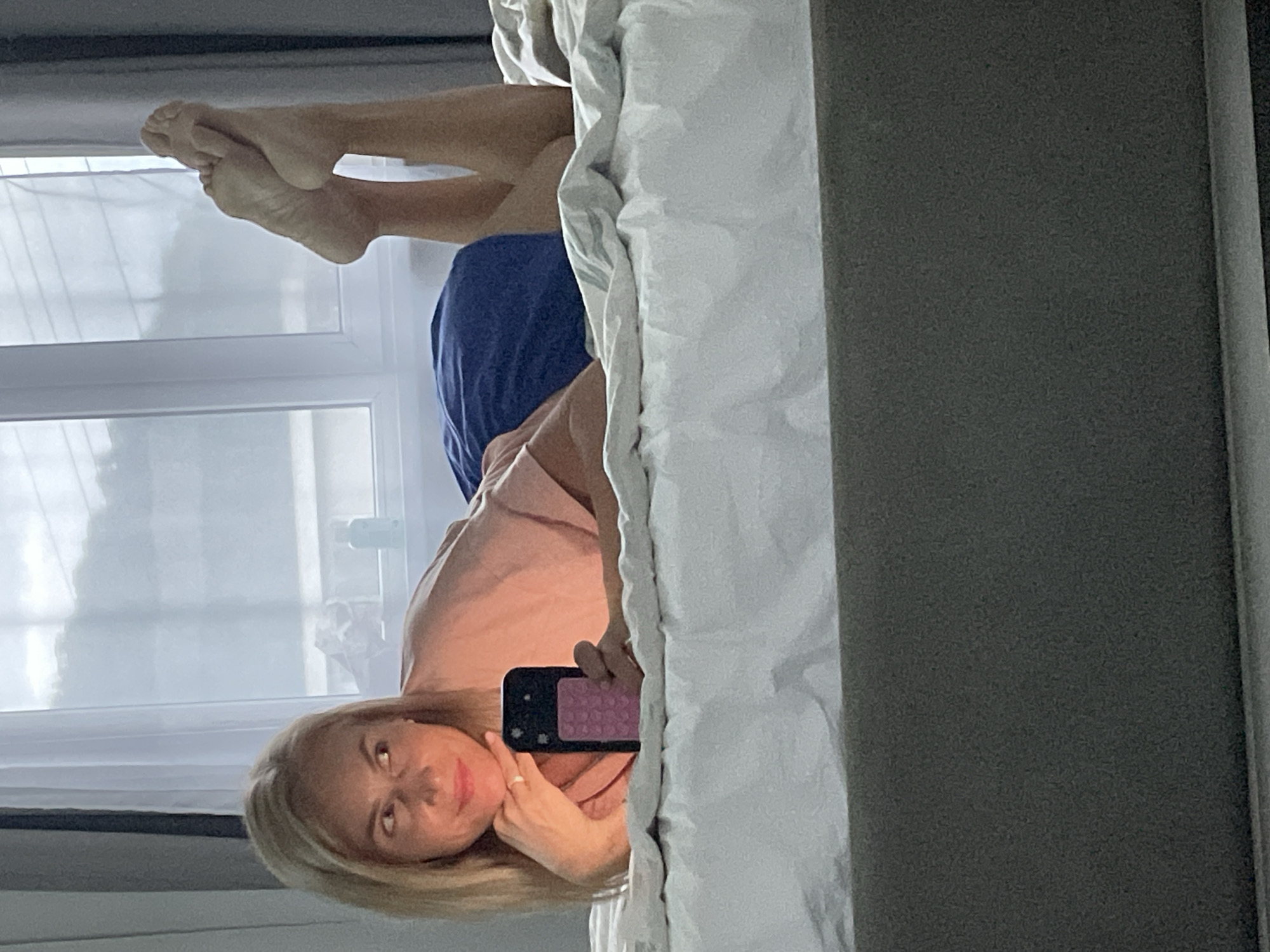 A blonde woman is lying on a bed, facing the viewer while holding a smartphone. Her feet are visible, extending toward a window with white curtains. She is wearing a pink shirt and blue bottoms. The room appears to be indoors, with a partially visible bed and wall. There is no indication of the location being Baranovka, Russia.