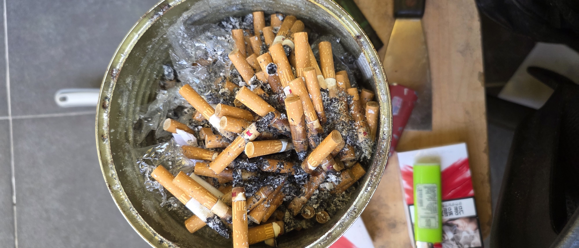 An overhead, close-up shot captures the interior of a large, round, metal receptacle filled with numerous cigarette butts. The butts are of various lengths and are predominantly orange and white, with some showing signs of ash and burns. The receptacle itself appears to be a repurposed tin can, with visible rust and wear on its rim. The surrounding environment suggests an outdoor setting, with a textured grey surface, possibly a tiled patio, visible in the upper portion of the image. A white object, potentially a handle or a light fixture, is positioned in the background. In the foreground, a green lighter rests on top of a pack of cigarettes, which features red and white graphics and Korean text. The overall scene is dimly lit, with the focus sharply on the collection of cigarette butts within the receptacle.
