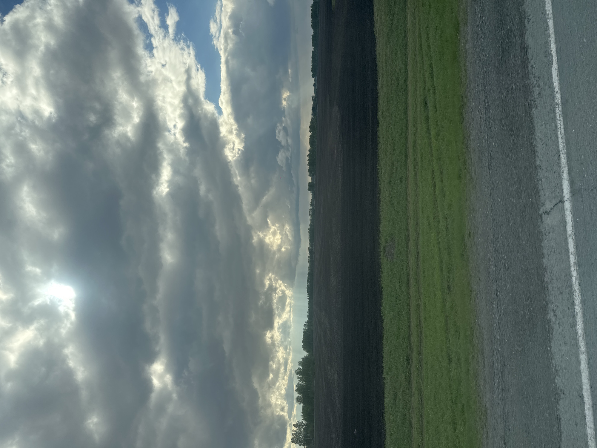 The image shows a landscape divided vertically into three distinct sections. The left side is dominated by a cloudy sky with sunlight filtering through the clouds. The middle ground appears to be a field of dark soil, likely recently plowed or tilled, separated from the sky by a line of trees. The right side consists of green vegetation adjacent to a grey asphalt road with a white line marking the lane.
There are no people visible. There is no location info visible in the image to be able to pinpoint the location in Yuzhnyi, Russia.