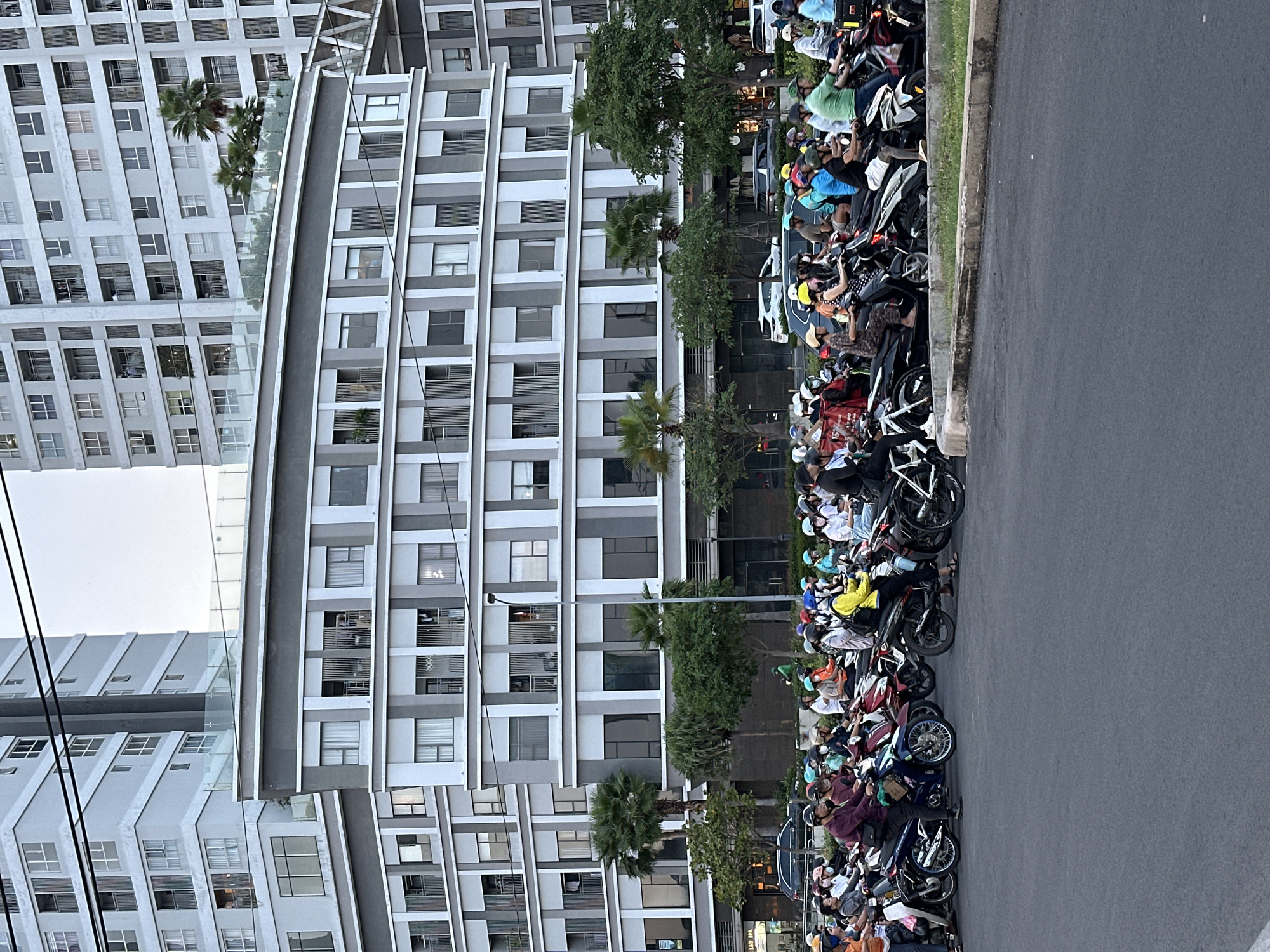The image shows a street scene in Thủ Đức, Vietnam. A row of parked motorbikes occupies the right side of the frame, with a large building on the left. Many people are sitting on the motorbikes, some wearing helmets. The building is multi-story with a repetitive window pattern. Some trees and landscaping are visible between the motorbikes and the building. The street appears to be paved. Overhead power lines are visible in the upper left.