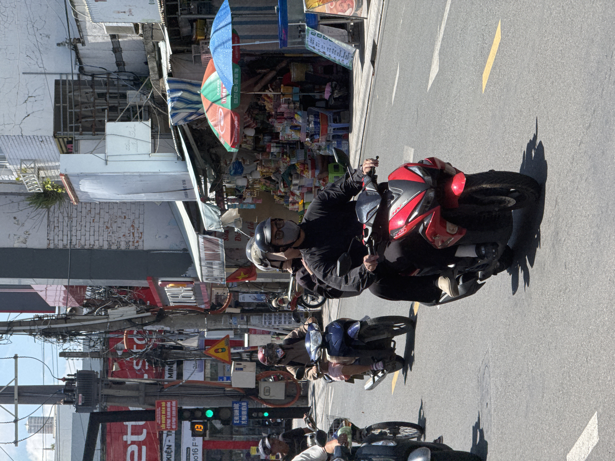 The image depicts a street scene in Phú Thủy, Vietnam, on a sunny day. Several individuals are riding motorbikes on a paved road. In the foreground, a person on a red motorbike wearing a black long-sleeved shirt, black pants, and a light-colored shoe is visible from the chest down. To their left, another person on a motorbike is partially visible. In the middle ground, a person wearing a brown jacket and helmet rides a blue motorbike, with another individual behind them also wearing a helmet. Further back, more motorbikes and pedestrians are present. The background features buildings with signs, utility poles with numerous wires, and an intersection with a traffic light showing green. Colorful umbrellas and displays of goods are set up along the roadside, suggesting a market or commercial area. The overall atmosphere is that of a busy urban environment.