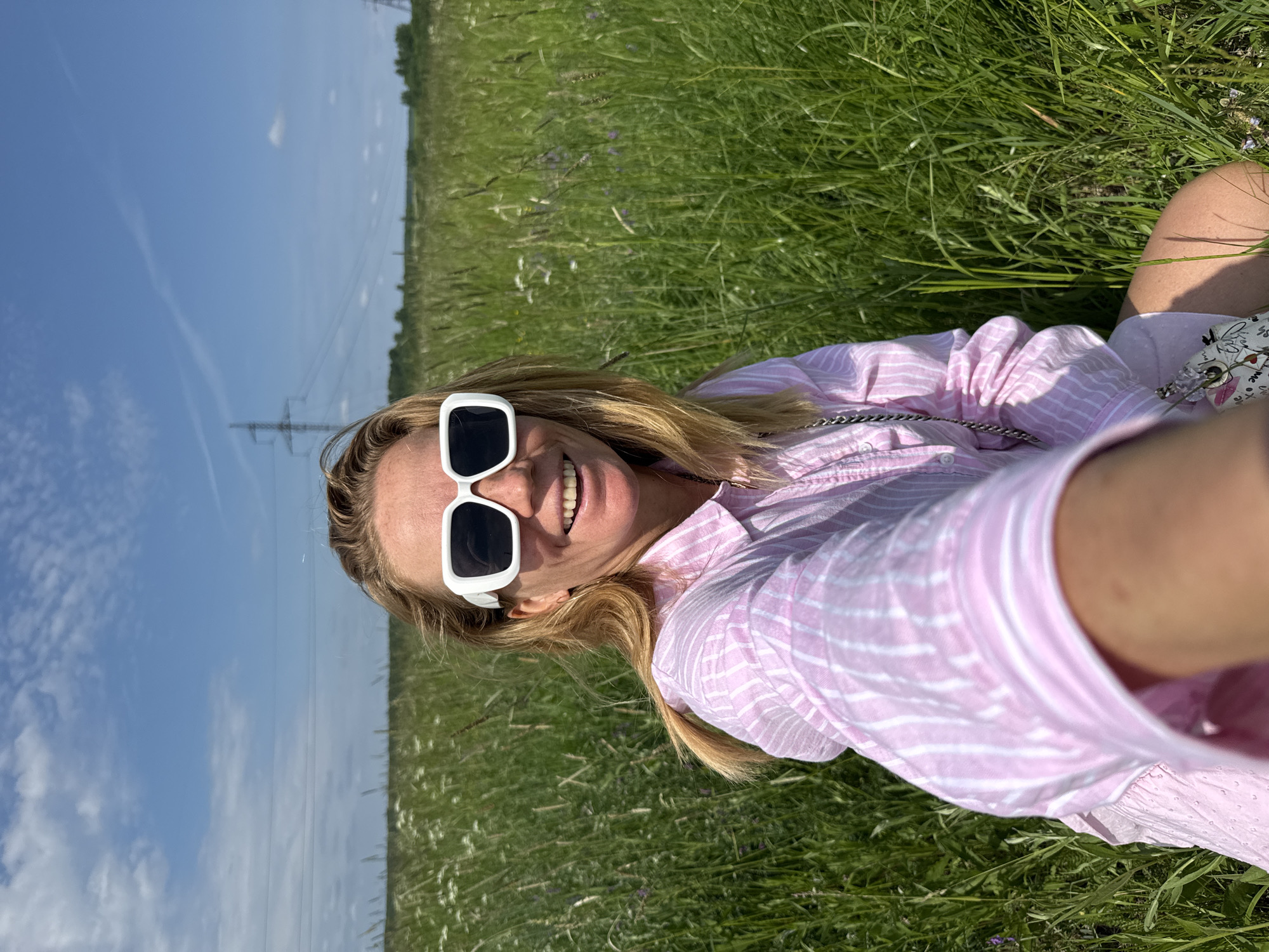 A blond woman with sunglasses and a pink striped shirt is sitting in a field of green grass under a blue sky with some clouds. There are power lines in the background. The woman is smiling and looking at the camera, holding the camera out to take a selfie. The location is described as Menevsha, Russia.