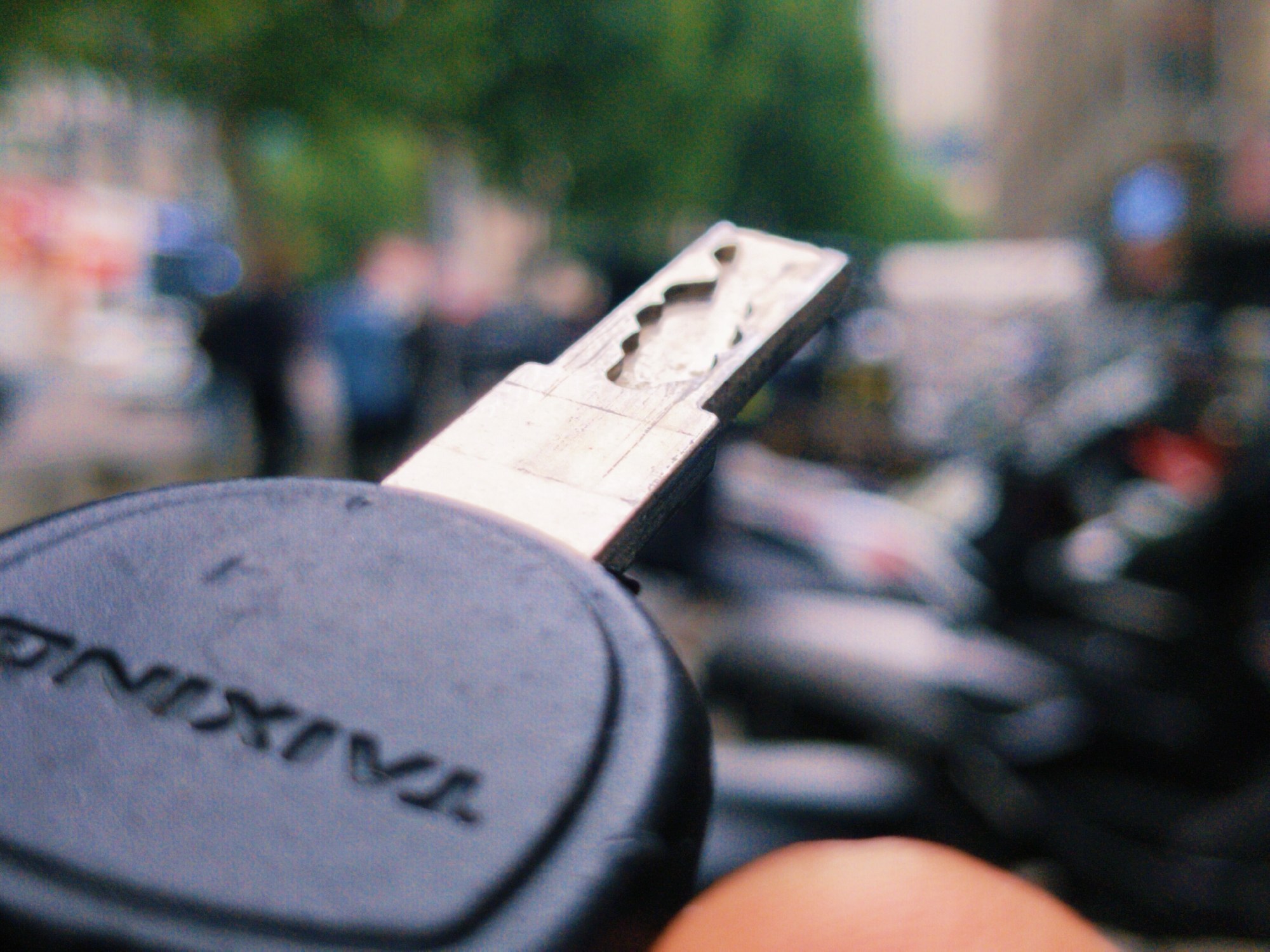 A close-up shot of a key with the word "TAIXING" on the black plastic handle. The key is held, and the background is blurry, showing a street scene with people, vehicles, and greenery. The location appears to be an urban area in daylight.