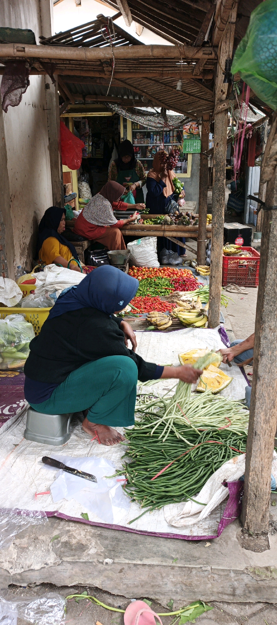 A scene unfolds at a roadside market stall in Kasiyan, Indonesia, featuring several vendors and a variety of fresh produce. In the foreground, a woman wearing a blue hijab and green pants is seated on a small gray stool, tending to a large pile of long green beans. Her bare feet are visible, and a knife lies on a piece of white plastic sheeting in front of her. Beside the beans, a cut piece of jackfruit is visible on the ground.
Behind her, to the left, another woman in a yellow top and blue hijab is seated, looking towards the camera with her tongue slightly out. A yellow plastic basket is behind her. Further back, a woman in a red top and a patterned headscarf is seated on a wooden bench, handling some vegetables. Another woman, also in a headscarf, stands behind her, wearing an apron and holding a green plastic bag. The stall itself is covered by a rustic bamboo and wood roof, with various goods displayed on shelves in the background, including bottled drinks and other packaged items. In front of the stall, rows of tomatoes and chilies are neatly arranged on the ground. Several bunches of bananas are also visible. To the right, a red crate is filled with more bananas, and a pair of flip-flops rests on the ground nearby. The overall environment suggests a bustling, open-air market setting.