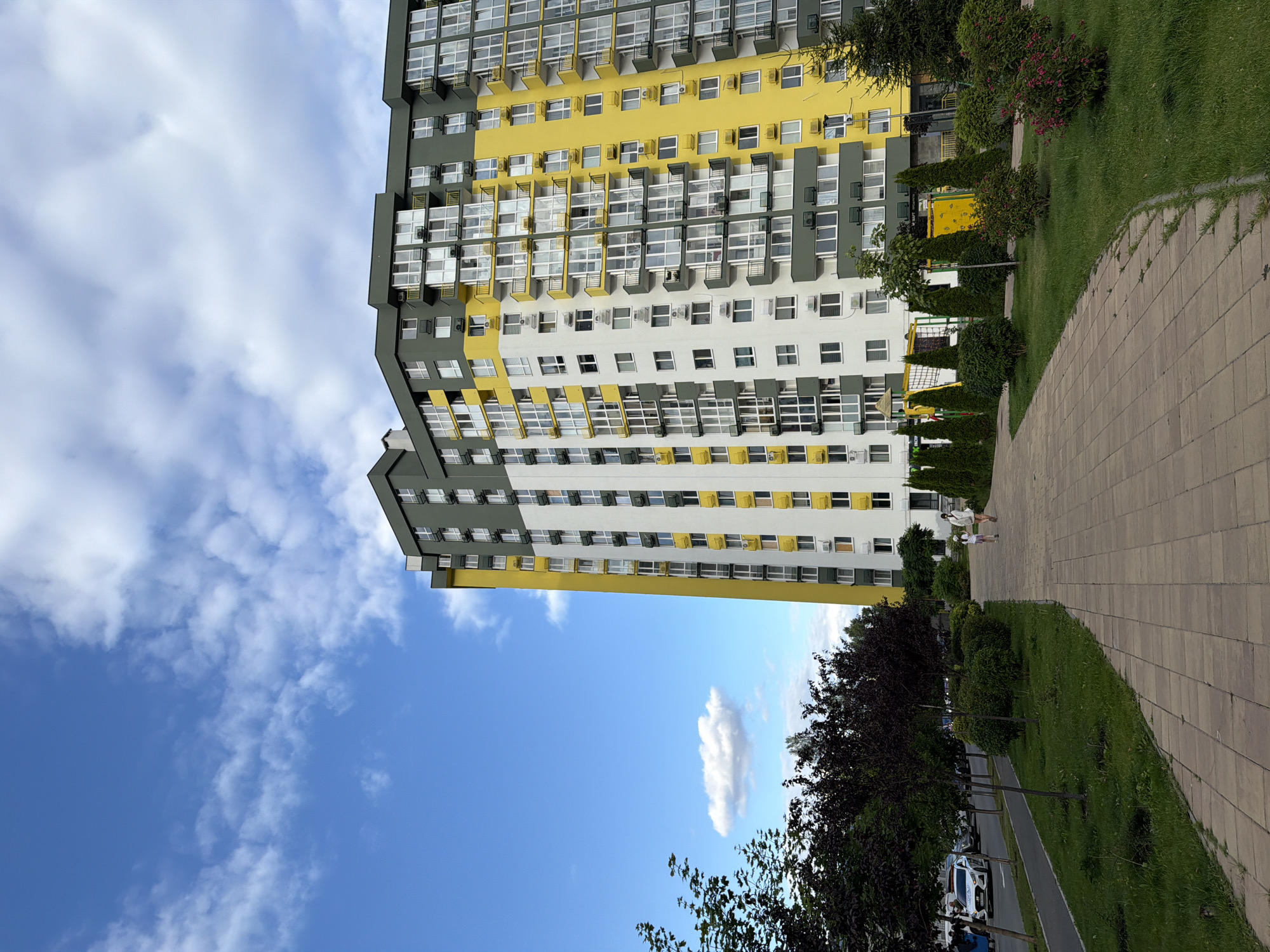 The image depicts a modern residential building with a yellow, white, and green facade, characteristic of contemporary urban architecture in Kyiv, Ukraine. The building is tall, with multiple floors featuring balconies and numerous windows. In the foreground, a paved walkway with beige square tiles leads through a well-maintained green area with trees and shrubs. Two individuals, appearing to be a woman and a child, are walking away from the camera on the walkway. To the left of the walkway, a street is visible with parked cars, including a white car with flashing lights, suggesting a police or emergency vehicle. The sky above is blue with scattered white clouds, indicating fair weather. The scene is set in a residential district, likely a newly developed area given the modern building and landscaping.