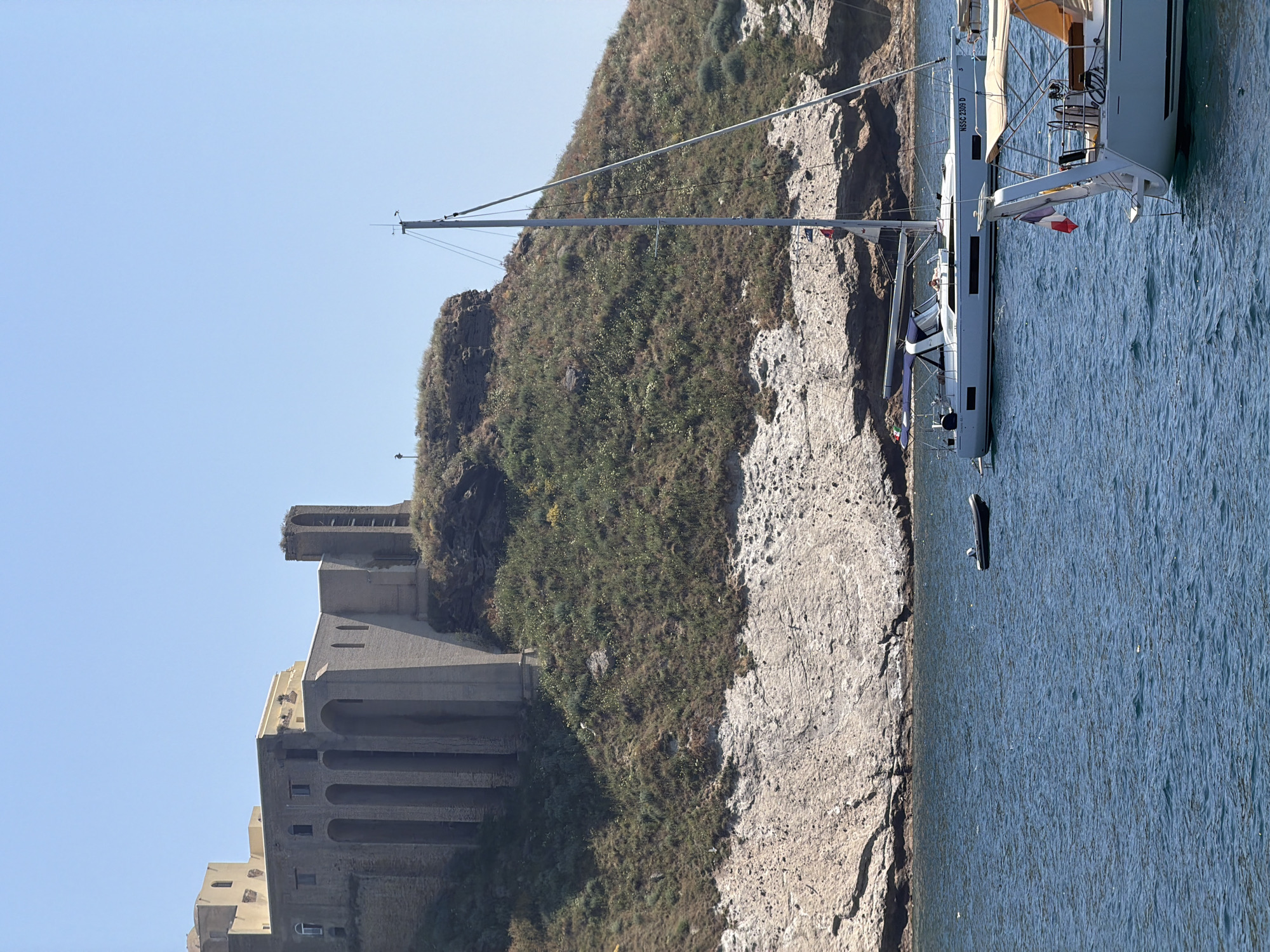 The image depicts a coastal scene with a sailboat docked next to a rocky coastline in Procida, Italy. The landmass rises to form a small hill covered in vegetation. Built into the hillside is a multi-tiered stone structure with arched supports, characteristic of the architecture in Procida. The sailboat is partially visible, with its mast extending high and its hull positioned in the water. The sea is calm and blue. The sky is clear and light blue. Overall, the image captures a typical coastal landscape of Procida, with its unique architecture integrated into the natural environment.