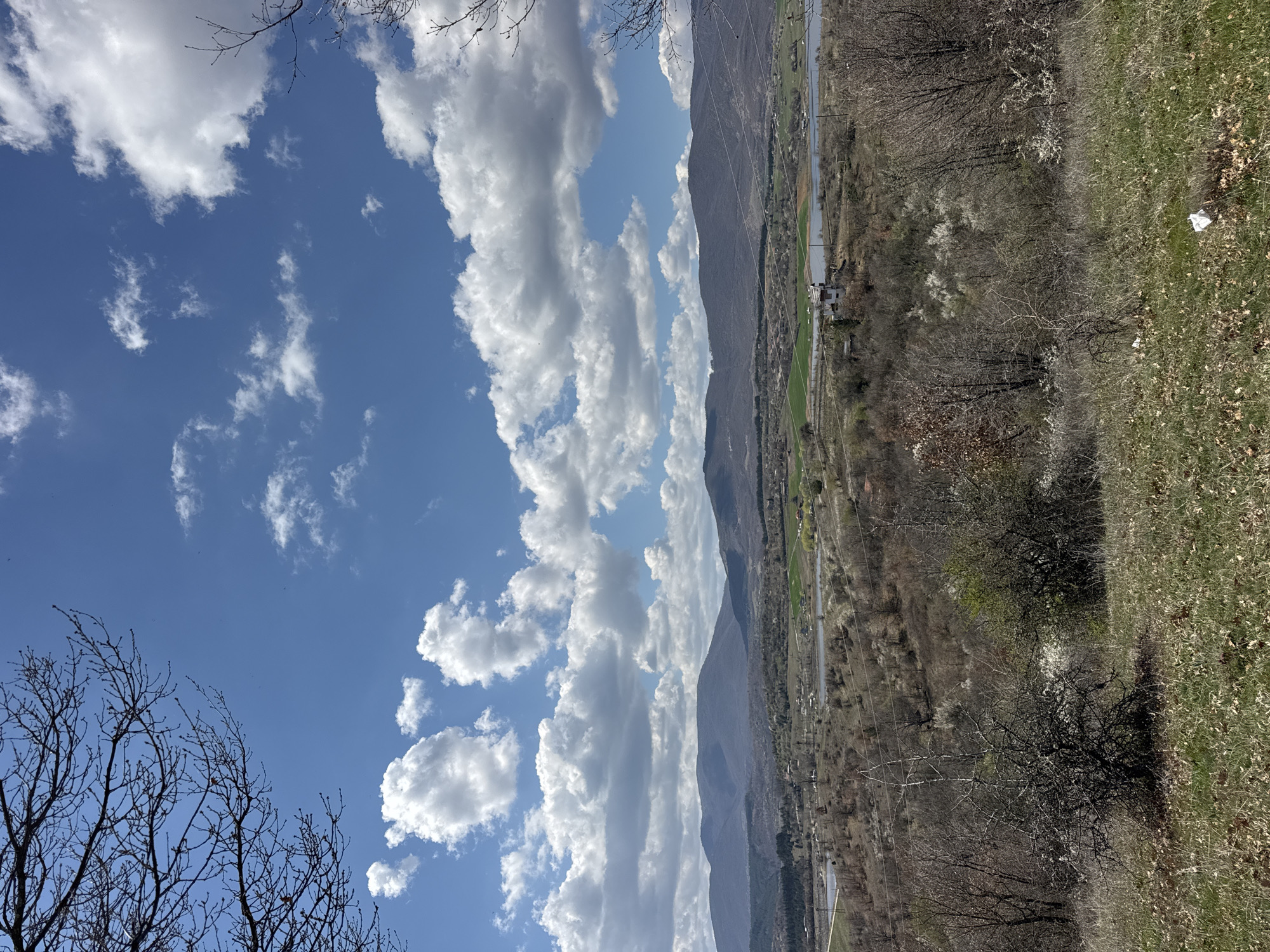 The image captures a landscape view in Logodaj, Bulgaria. The scene is divided between a clear blue sky with scattered cumulus clouds and a mountainous terrain. A mountain range runs vertically through the center of the image. To the right of the mountains, a valley is visible, featuring a river or stream, patches of green fields, and sparse buildings. The foreground on the right shows a grassy slope mixed with bare trees and foliage. The sky on the left displays branches of trees.